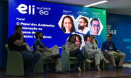 Cinco palestrantes estão sentados em poltronas verdes alinhadas em um palco de madeira durante o evento "Eli Summit GO". Ao fundo, um grande painel digital azul e verde exibe fotos de perfil e os nomes dos debatedores, além do título "O Papel dos Ambientes de Inovação no Sucesso de Negócios Inovadores". No centro do palco, uma mulher fala ao microfone enquanto os demais integrantes, compostos por três homens e outra mulher, escutam atentamente.