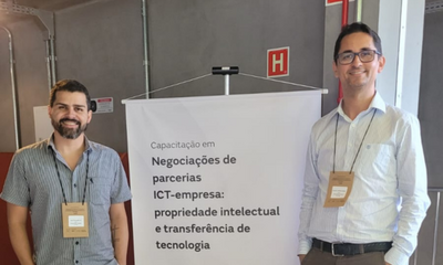 Dois homens sorridentes, usando crachás de identificação, posam ao lado de um banner branco em um ambiente interno com paredes cinzas. O banner anuncia uma capacitação em "Negociações de parcerias ICT-empresa", focada em propriedade intelectual e transferência de tecnologia. O homem à esquerda veste uma camisa cinza de mangas curtas, enquanto o da direita usa uma camisa social clara e óculos.