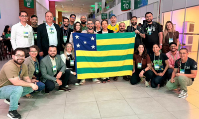 Um grupo numeroso de pessoas posa sorridente em um amplo corredor interno de eventos, posicionado em torno da bandeira do estado de Goiás, que é segurada aberta ao centro. Os participantes, homens e mulheres vestindo roupas casuais e sociais, utilizam crachás oficiais pendurados no pescoço. Ao fundo, identificam-se placas de sinalização de setores e salões com números, além de estruturas de estandes e iluminação clara.