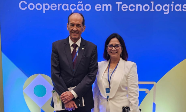 Um homem vestindo terno escuro e uma mulher em um blazer branco posam sorridentes à frente de um painel azul onde se lê "Cooperação em Tecnologias". Ambos utilizam crachás institucionais com cordões azuis e seguram materiais de apoio, como pastas ou cadernos. A imagem registra a participação de representantes em um evento oficial, transmitindo uma atmosfera de formalidade e parceria profissional.