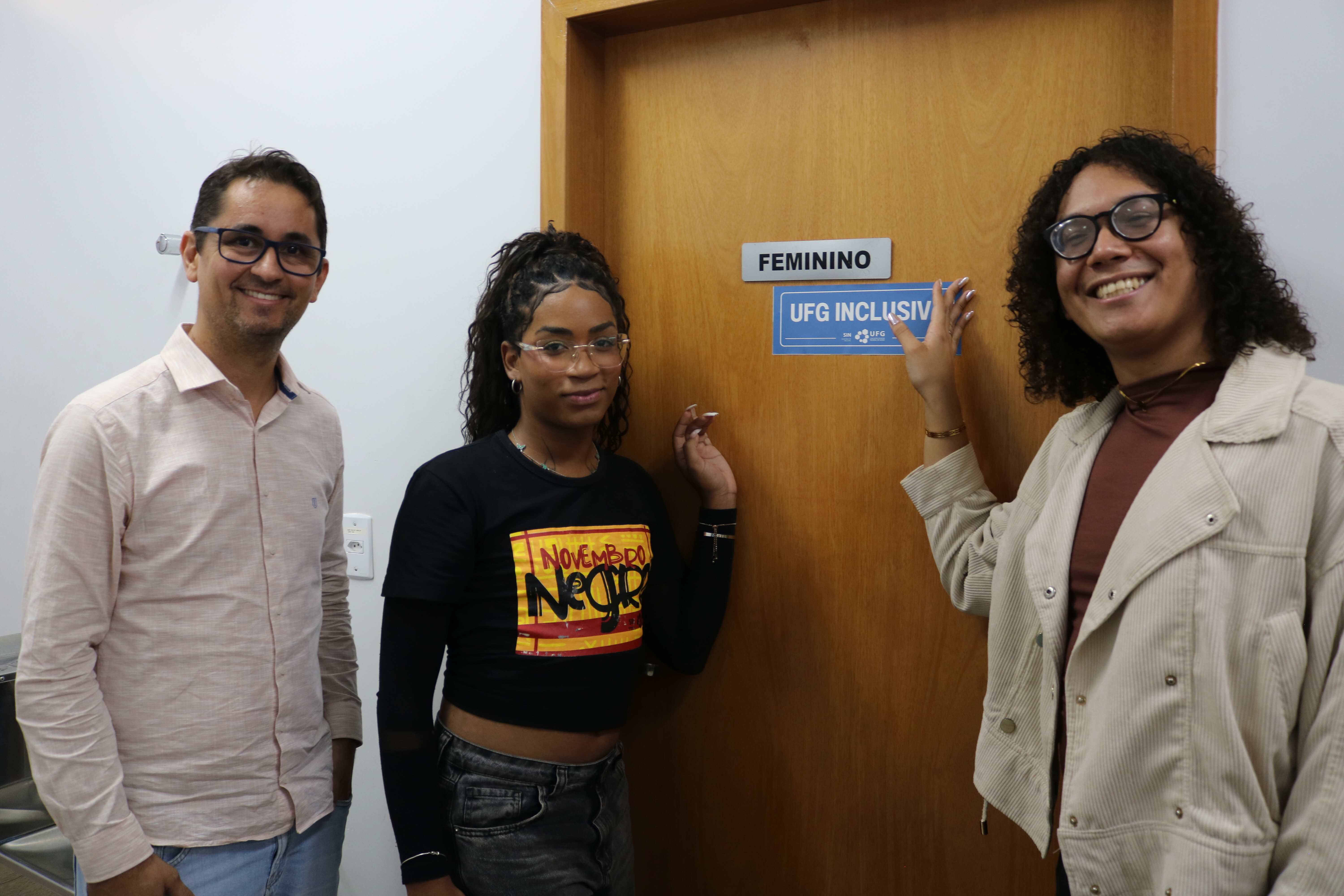 Três pessoas sorridentes — um homem e duas mulheres — posam em um corredor ao lado de uma porta de madeira. Uma das mulheres aponta para o adesivo azul "UFG INCLUSIVA" fixado logo abaixo da placa de identificação "FEMININO". O registro captura o grupo em um plano médio, destacando a ação de sinalização inclusiva no ambiente da universidade.