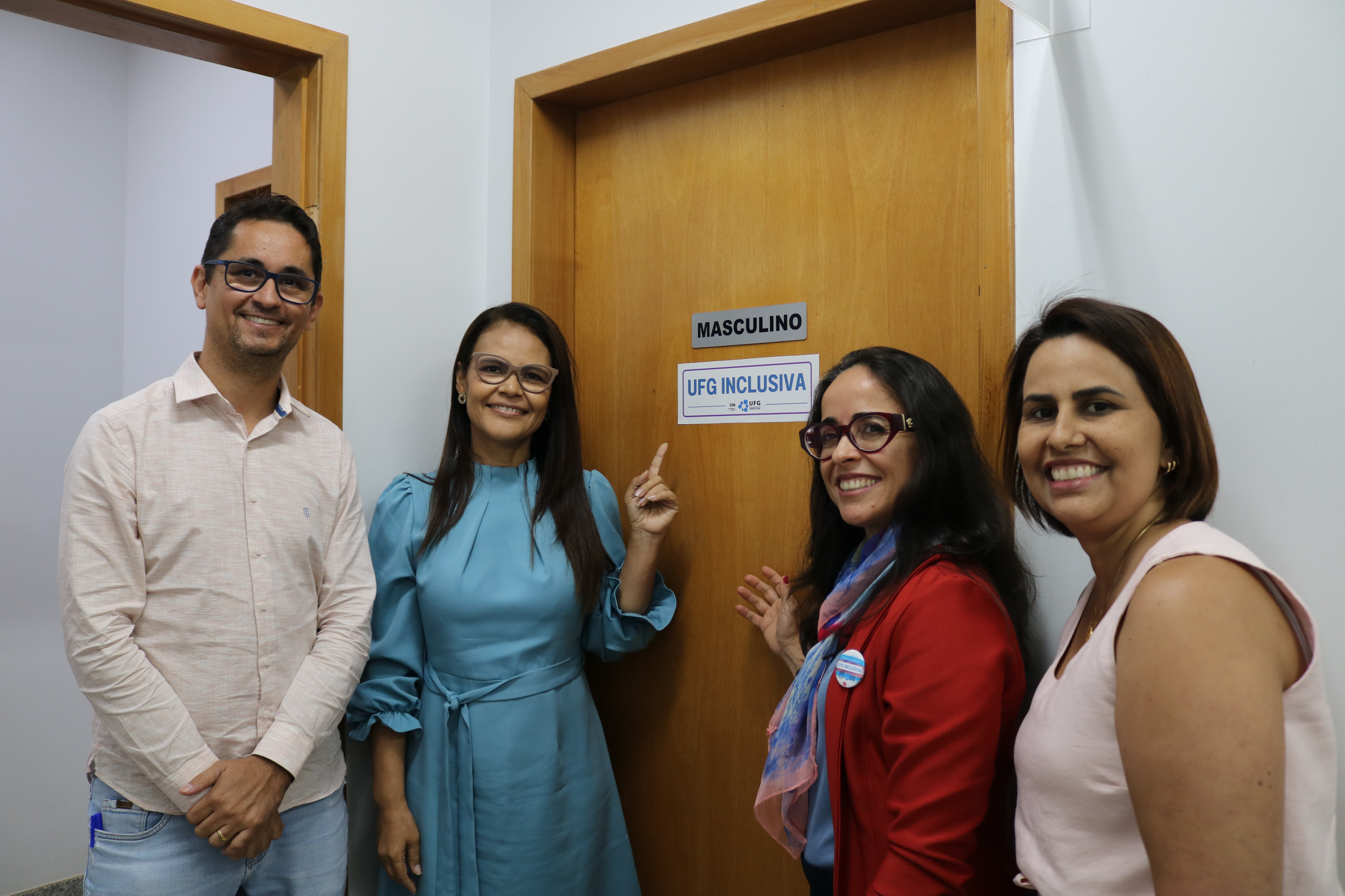 Quatro pessoas sorridentes, um homem e três mulheres, posam para a fotografia em um corredor institucional ao lado de uma porta de madeira. A mulher de vestido azul aponta para o adesivo "UFG INCLUSIVA" fixado abaixo da placa "MASCULINO", celebrando a sinalização inclusiva. O grupo está alinhado verticalmente em relação à câmera, transmitindo uma atmosfera de realização e parceria.