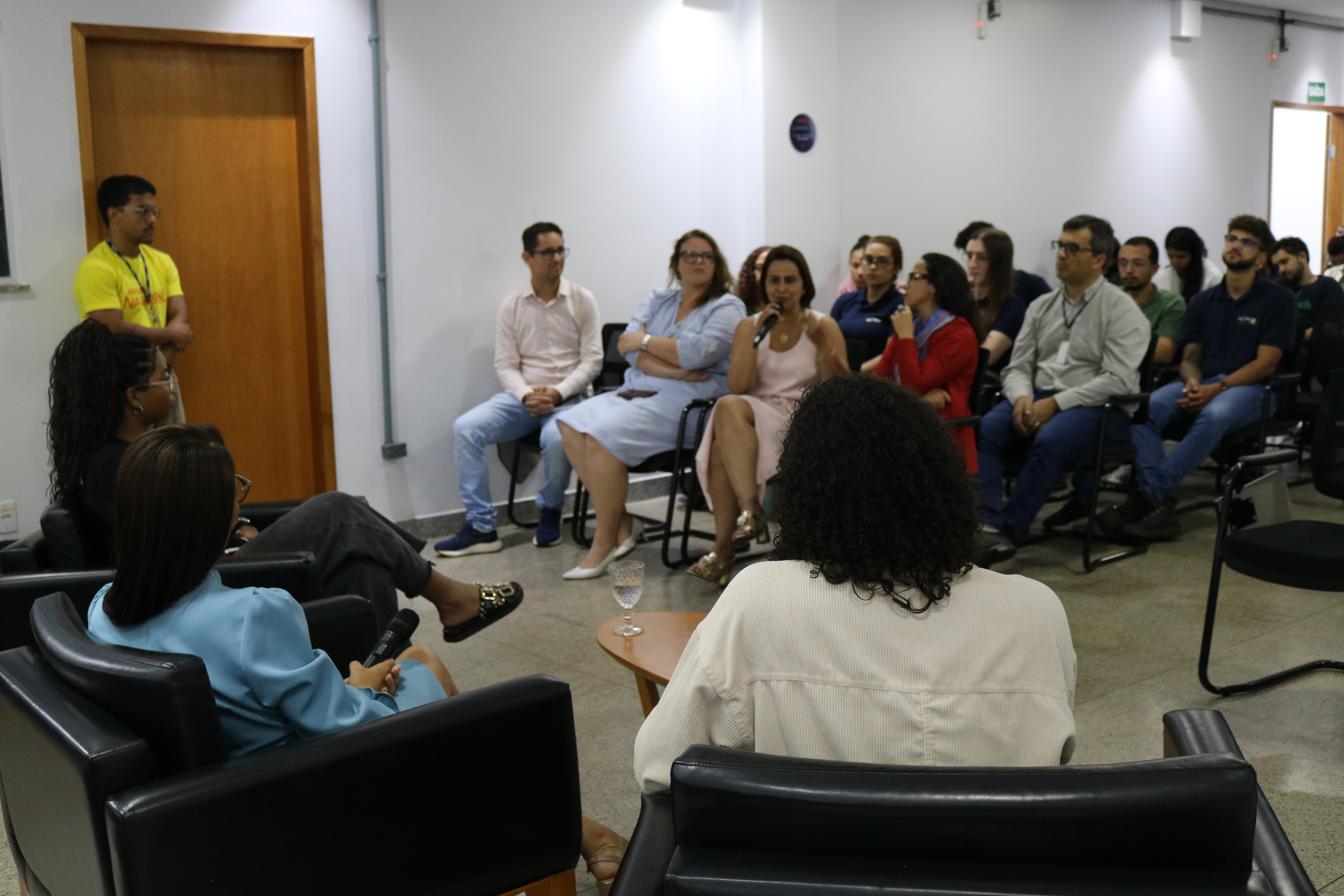 A imagem mostra uma mulher na plateia falando ao microfone para o público presente. Em primeiro plano, aparecem as costas de participantes sentados em poltronas pretas, voltados para a audiência. O grupo acompanha o debate atentamente em um ambiente institucional bem iluminado.