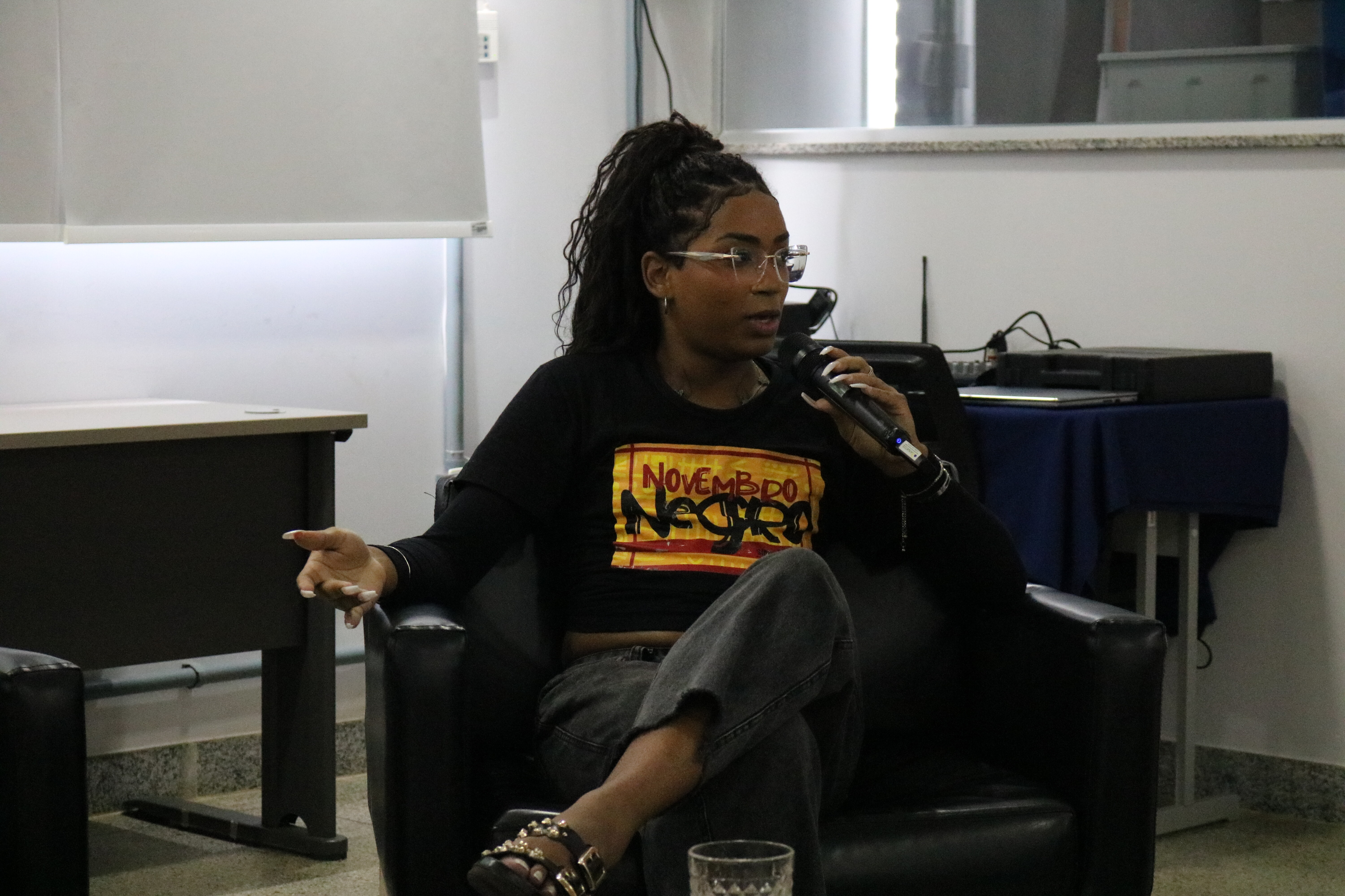 Uma mulher de cabelos longos presos e óculos fala ao microfone enquanto gesticula, sentada em uma poltrona preta. Ela veste uma camiseta preta com a estampa "NOVEMBRO NEGRO" em um ambiente de debate institucional. A cena registra um momento de fala ativa e engajamento durante o evento realizado em local fechado.