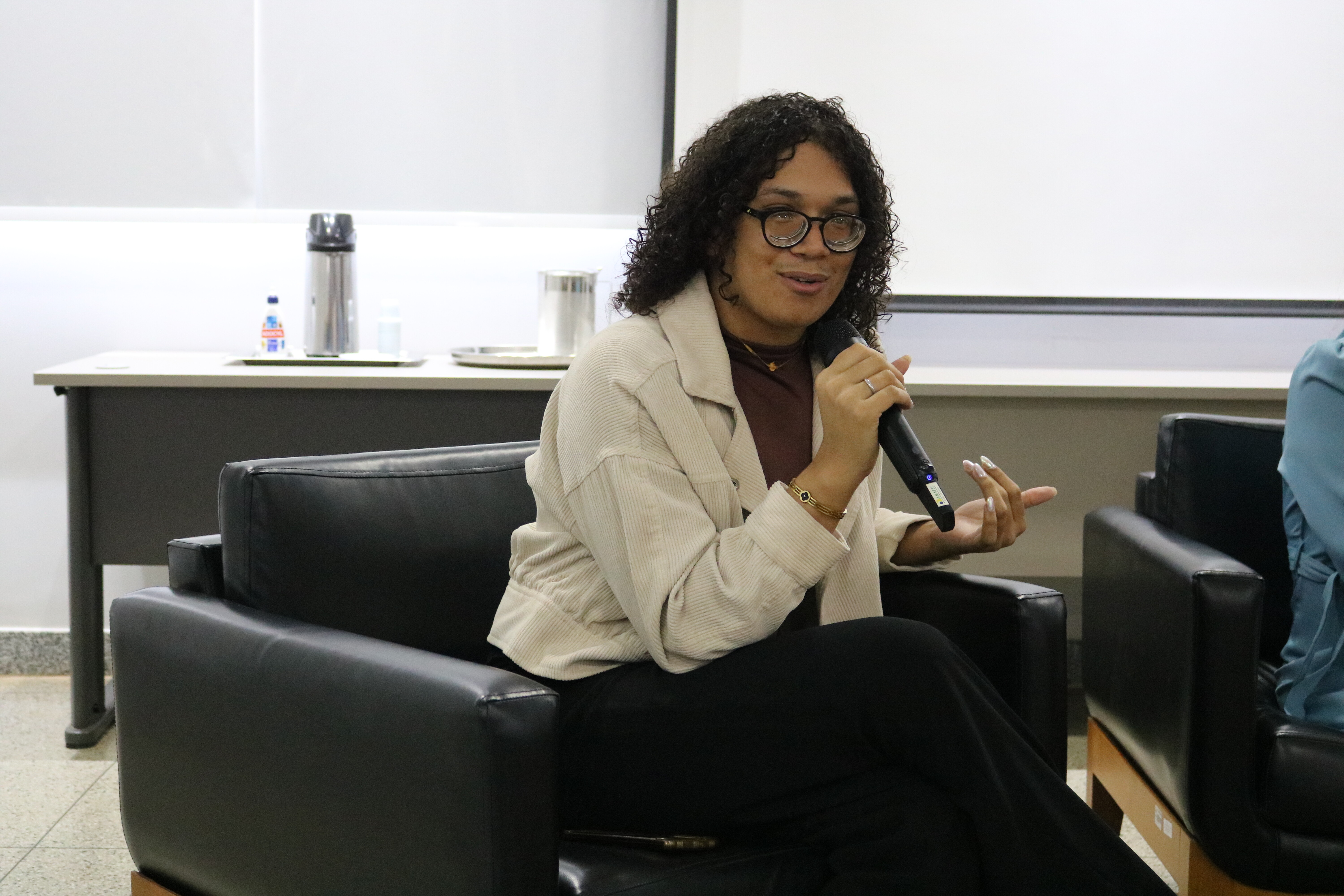 A pessoa de cabelos cacheados e óculos está sentada em uma poltrona preta enquanto fala ao microfone e gesticula. Ela veste uma jaqueta clara sobre roupa escura em um ambiente de palestra institucional. Ao fundo, observa-se uma mesa com garrafa térmica e uma tela branca de projeção.