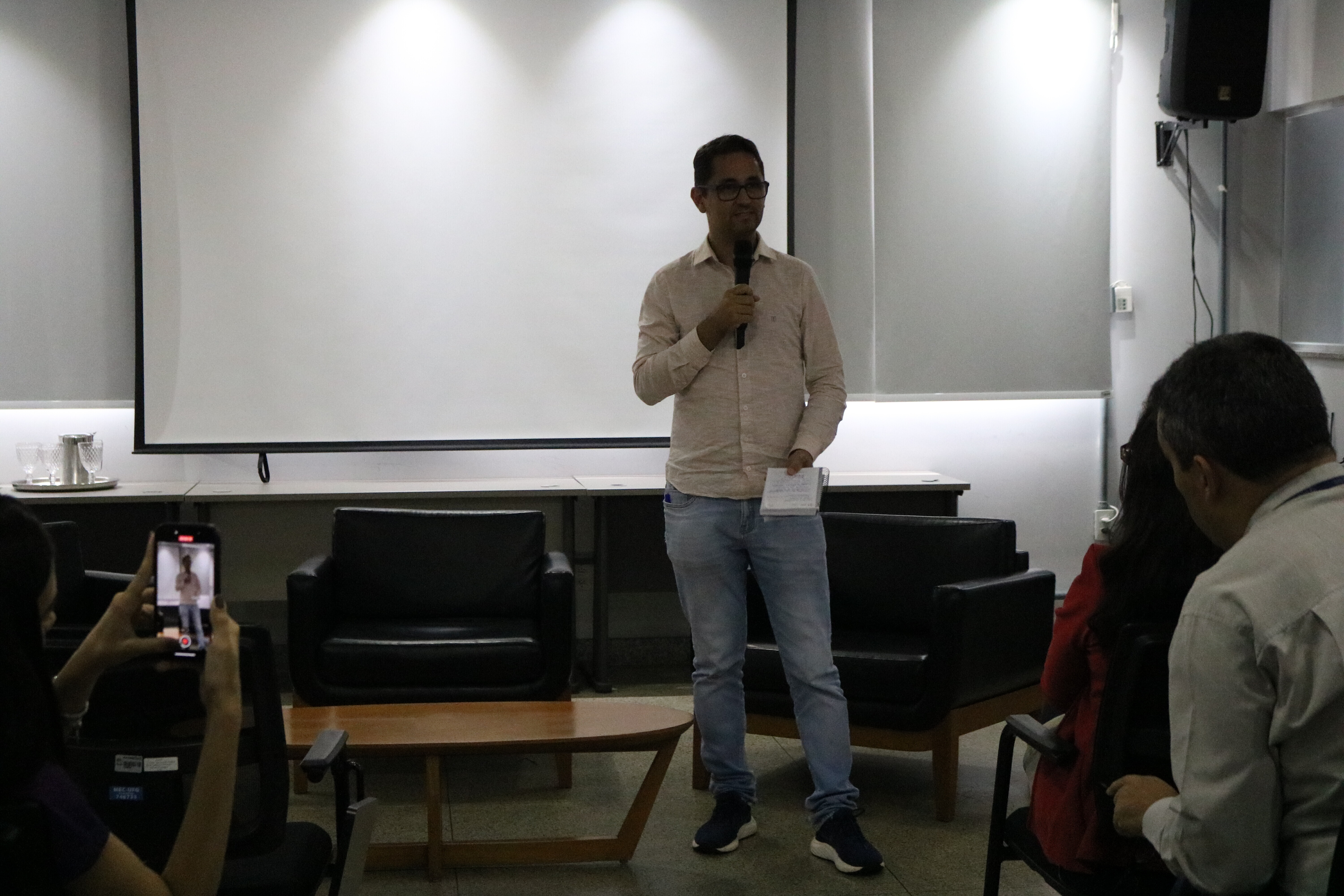 O diretor do PTS/UFG, Luizmar Adriano, segura um microfone e um caderno enquanto fala em uma sala equipada com uma tela de projeção branca ao fundo. Ele veste uma camisa clara e calça jeans, posicionado entre poltronas pretas e uma mesa de centro de madeira. À esquerda, em primeiro plano, uma pessoa registra a apresentação utilizando um smartphone.