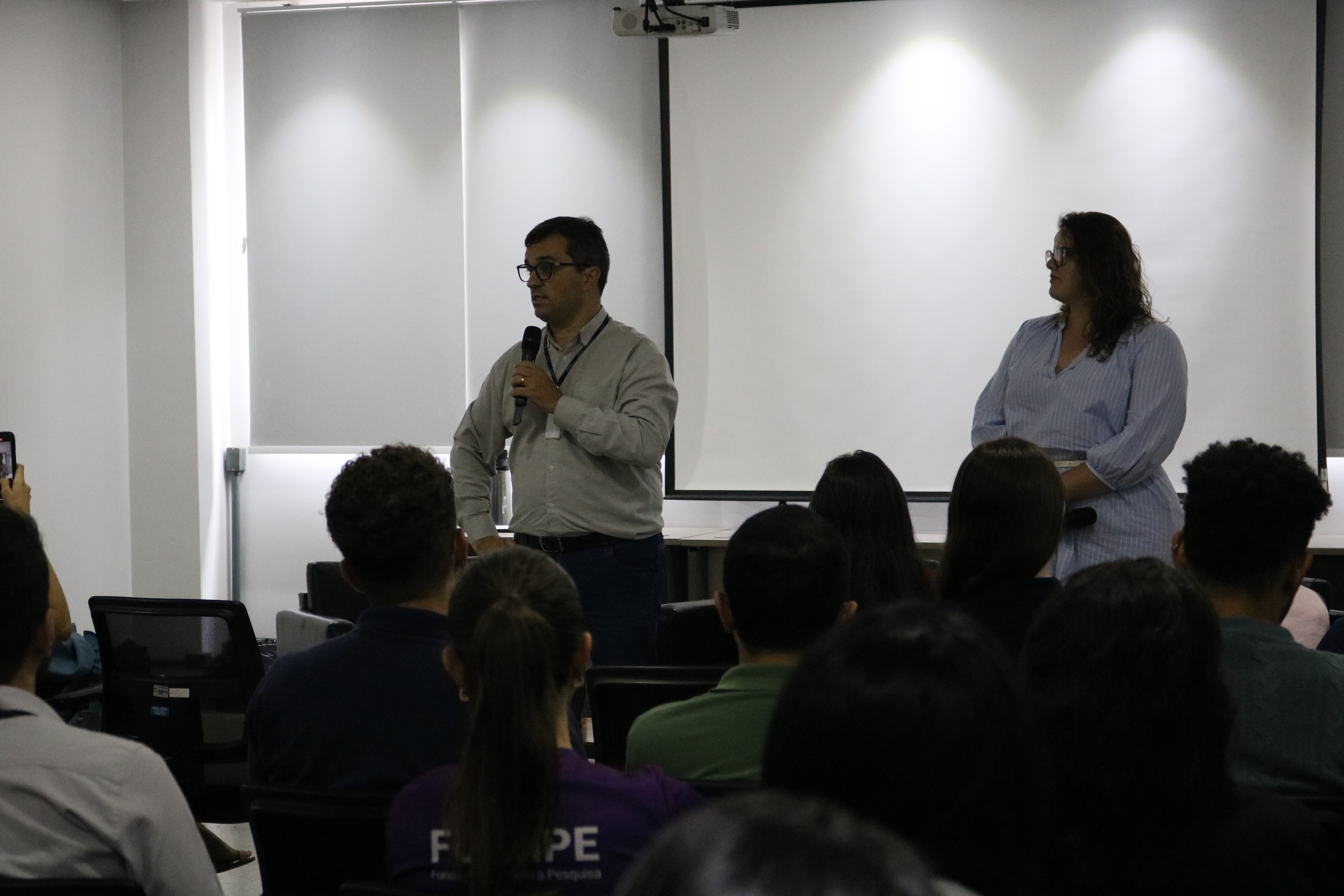 Um homem e uma mulher estão de pé à frente de uma plateia em uma sala de palestras iluminada. O homem utiliza um microfone para falar, enquanto a mulher, vestindo uma blusa azul listrada, permanece ao seu lado diante de um telão branco. A imagem, capturada de trás dos espectadores, registra um momento de interação institucional com o público presente.