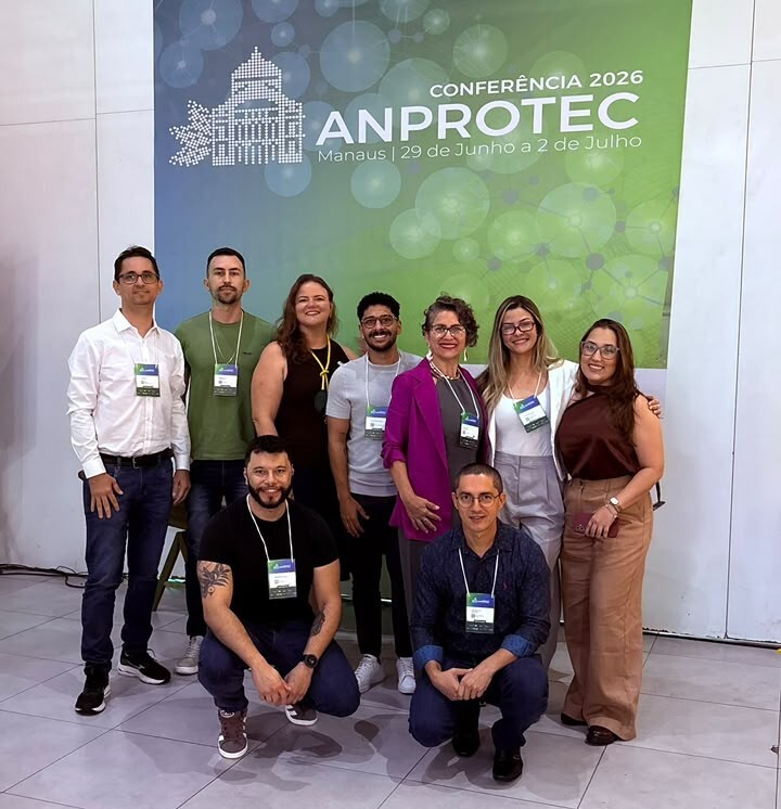 Um grupo de nove pessoas posa sorridente em frente a um grande painel azul e verde da "Conferência Anprotec 2026", realizada em Manaus. Os integrantes, vestidos com roupas casuais e sociais, utilizam crachás de identificação do evento pendurados no pescoço. No registro, sete pessoas aparecem posicionadas de pé, enquanto outras duas estão agachadas à frente sobre o piso claro
