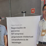 Dois homens sorridentes, usando crachás de identificação, posam ao lado de um banner branco em um ambiente interno com paredes cinzas. O banner anuncia uma capacitação em "Negociações de parcerias ICT-empresa", focada em propriedade intelectual e transferência de tecnologia. O homem à esquerda veste uma camisa cinza de mangas curtas, enquanto o da direita usa uma camisa social clara e óculos.