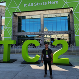 Um homem posa sorridente em frente ao San Jose McEnery Convention Center, onde se lê "NVIDIA GTC" e a frase "It All Starts Here" em um grande painel verde. À sua frente, destacam-se grandes letras e números verdes que formam a sigla "GTC 26". O cenário externo é amplo e moderno, sob a luz do dia, indicando a participação em um evento tecnológico de grande porte.