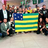 Um grupo numeroso de pessoas posa sorridente em um amplo corredor interno de eventos, posicionado em torno da bandeira do estado de Goiás, que é segurada aberta ao centro. Os participantes, homens e mulheres vestindo roupas casuais e sociais, utilizam crachás oficiais pendurados no pescoço. Ao fundo, identificam-se placas de sinalização de setores e salões com números, além de estruturas de estandes e iluminação clara.