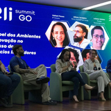 Cinco palestrantes estão sentados em poltronas verdes alinhadas em um palco de madeira durante o evento "Eli Summit GO". Ao fundo, um grande painel digital azul e verde exibe fotos de perfil e os nomes dos debatedores, além do título "O Papel dos Ambientes de Inovação no Sucesso de Negócios Inovadores". No centro do palco, uma mulher fala ao microfone enquanto os demais integrantes, compostos por três homens e outra mulher, escutam atentamente.