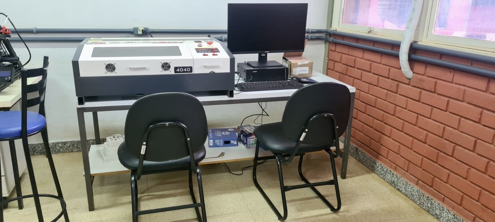 Na imagem há uma máquina de corte a laser portátil sobre uma bancada. Ao lado, na mesma bancada, há um monitor, teclado e computador. À frente, no chão, há duas cadeiras.