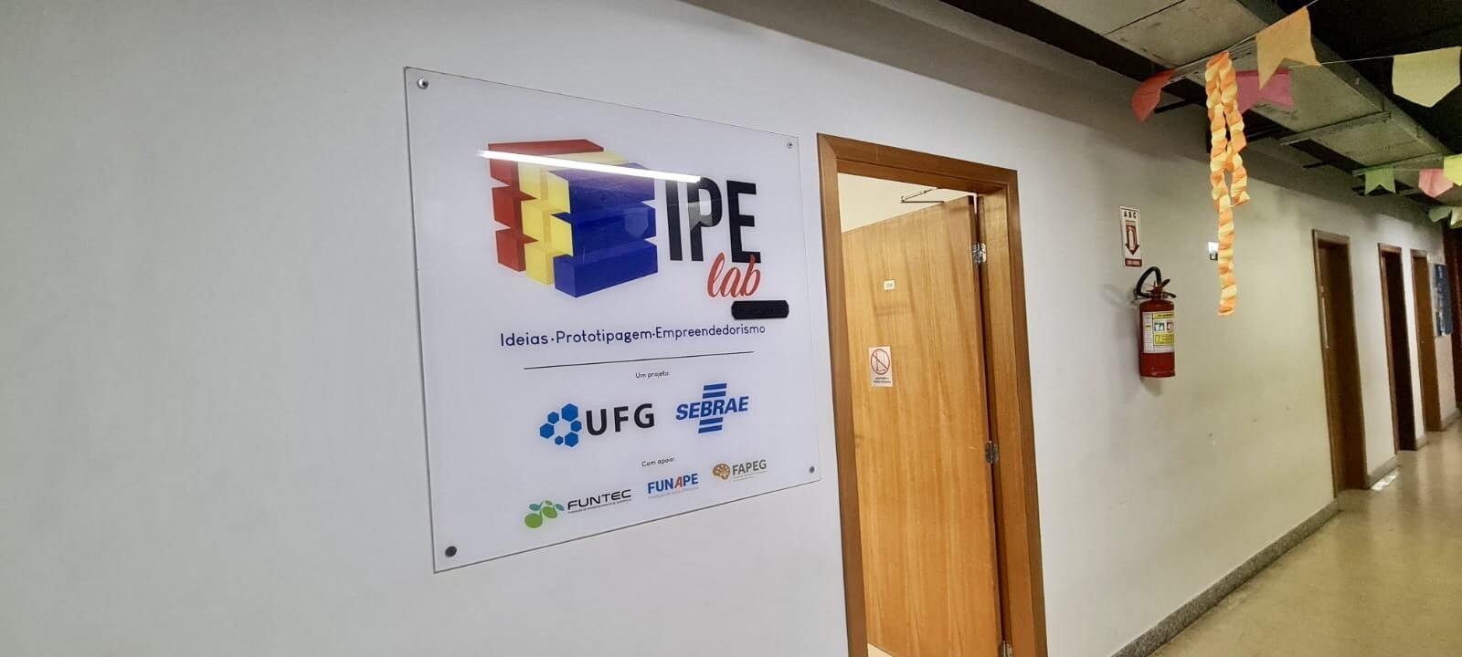 Foto da porta de entrada da unidade IPElab Física. Pode-se identificar na foto, um corredor com algumas portas, com foco na porta da sala do IPElab - Física, com uma placa ao lado identificando o laboratório com logotipo do IPElab e dos parceiros.