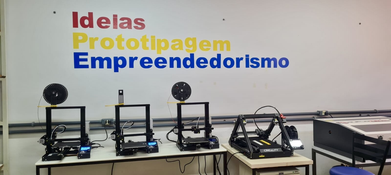 Foto impressoras 3D IPElab-Física. Na foto há uma parede com adesivos com as palavras: "Ideias", "Prototipagem" e "Empreendedorismo", em vermelho, amarelo e azul escuro, respectivamente. À frente da parede há uma bancada com 4 impressoras 3D FDM. No canto direito é possível identificar que há uma máquina de corte a laser portátil.