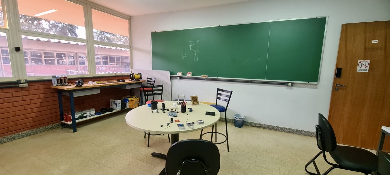 Foto da sala do IPElab-Física. Há uma mesa, uma bancada com alguns equipamentos, cadeiras e um quadro/lousa verde na parede.