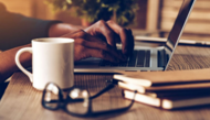 Fotografia mostra pessoa digitando em um notebook sobre uma mesa, cercado de materiais de escritório.