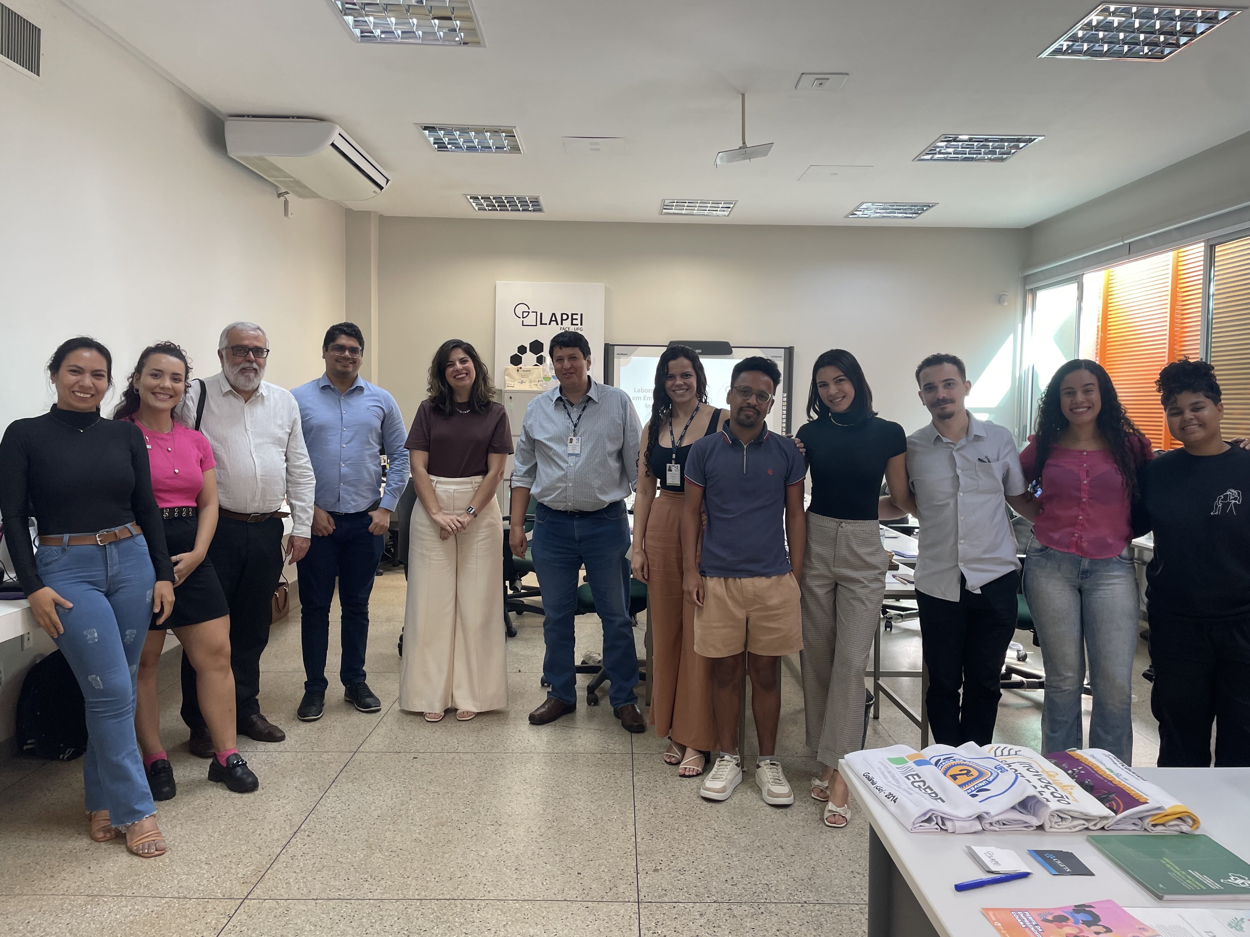 A foto mostra um grupo de 13 pessoas em pé, posando para a câmera dentro de uma sala bem iluminada, com janelas à direita. A maioria sorri. No fundo há um banner com a palavra "LAPEI", indicando que estão em um ambiente acadêmico ou profissional. As pessoas estão vestidas de forma variada, desde roupas sociais até trajes casuais. Na frente, sobre uma mesa, há panfletos, camisetas e materiais impressos. O ambiente é organizado, com mesas, cadeiras e equipamentos de escritório.