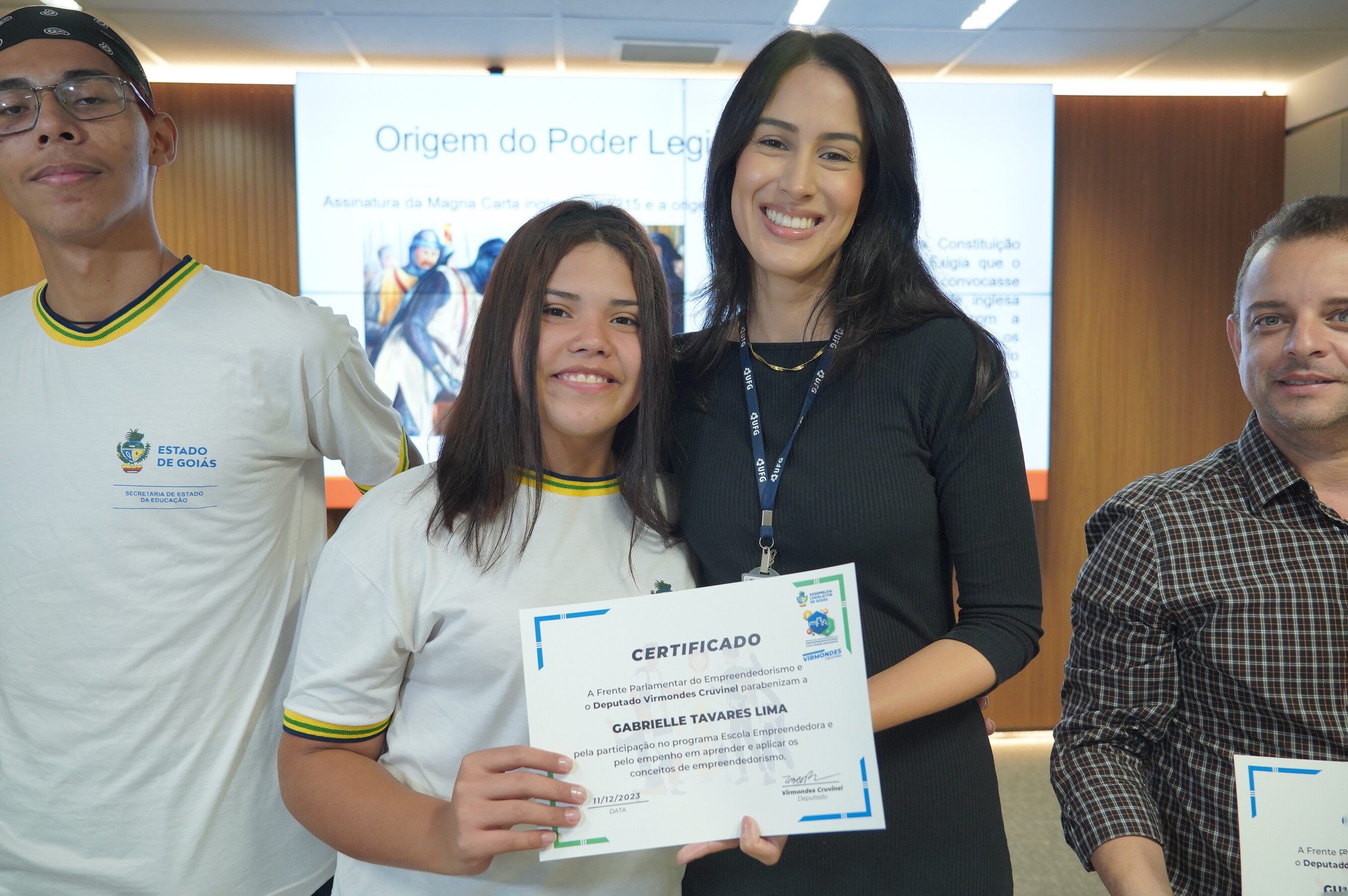 Aluna do CEPI Arco-Íris recebe homenagem. Na foto: uma mulher jovem, alta, morena, de vestido preto, entrega o certificado a uma aluna do ensino médio, morena, vestindo uniforme. 