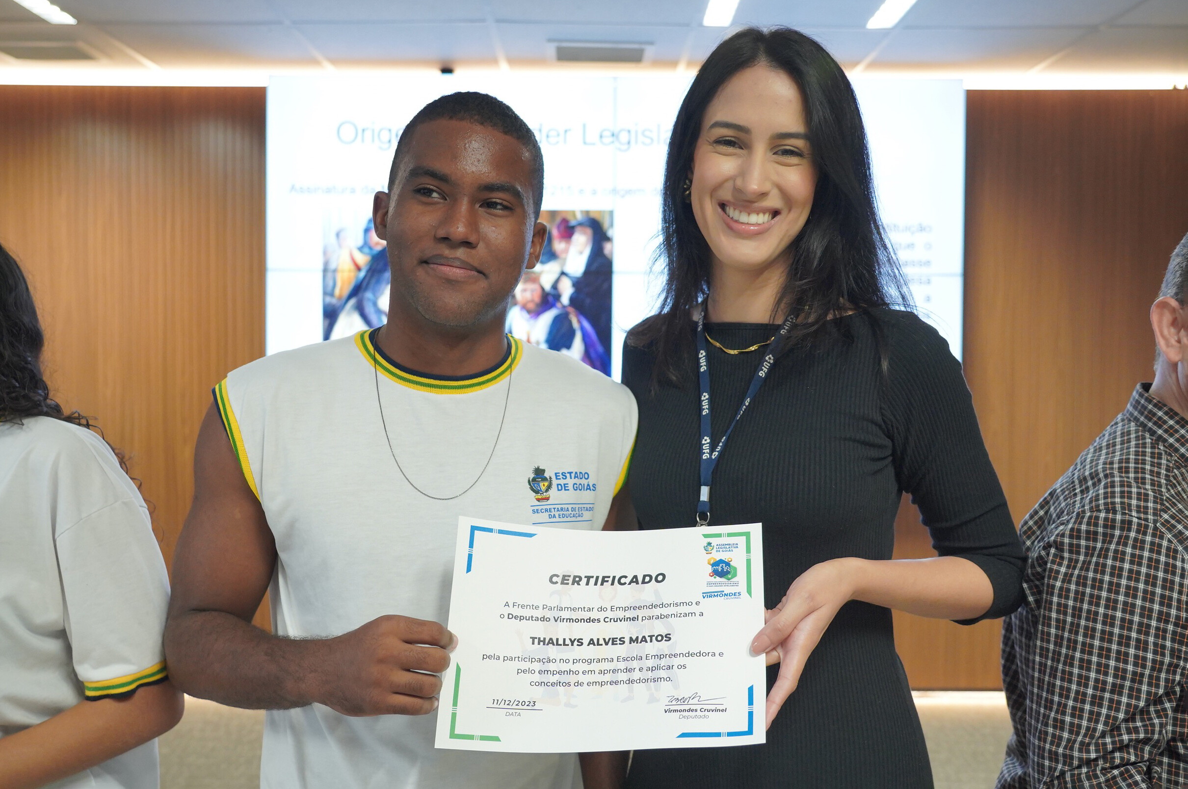 Aluno do CEPI Arco-Íris recebe homenagem. Na foto: uma mulher jovem, alta, morena, de vestido preto, entrega o certificado a um aluno do ensino médio, preto, vestindo uniforme. 