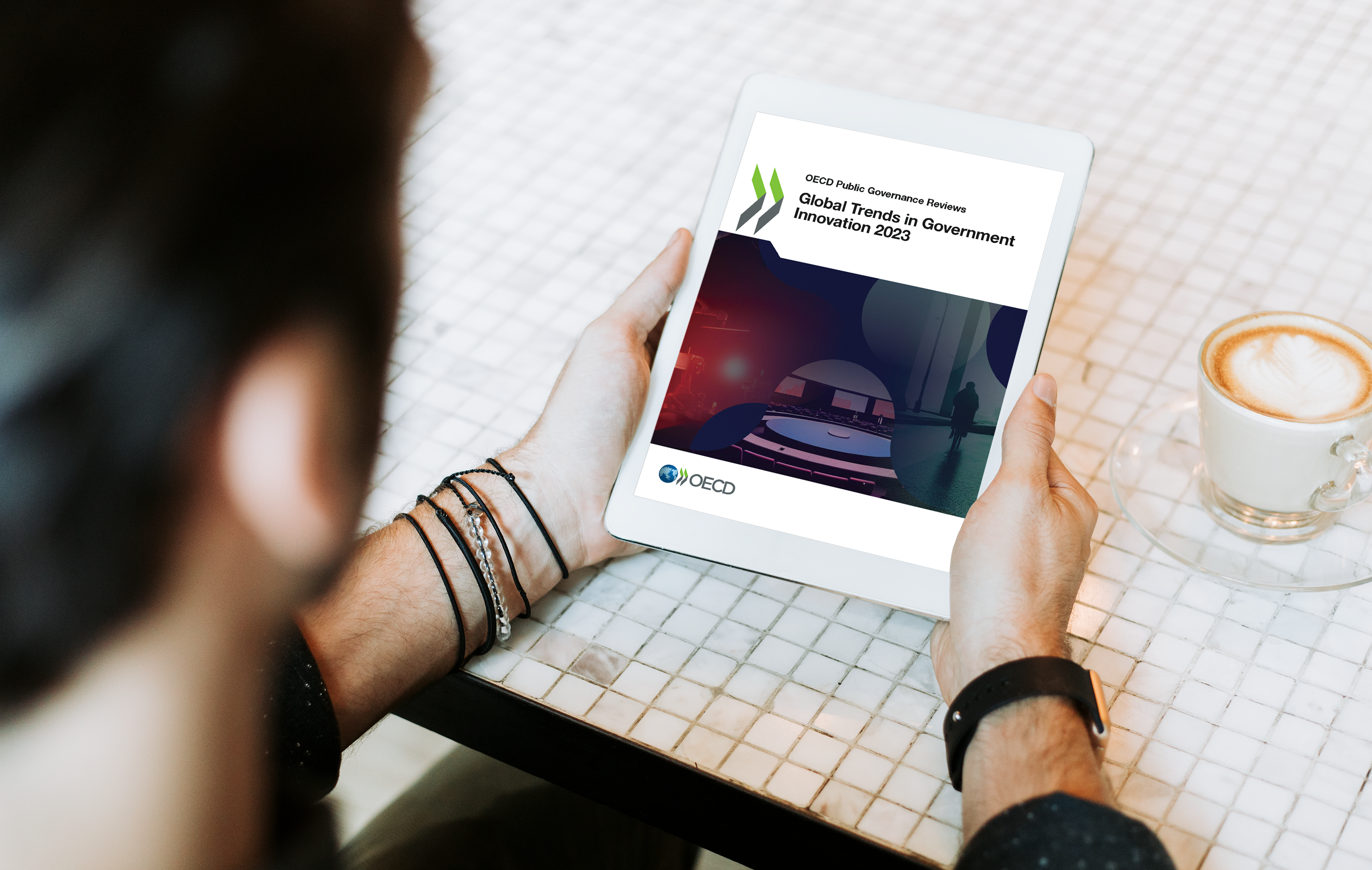 Foto corpo da notícia relatório OCDE. Homem de costas com um tablet na mão, com a imagem do relatório aberto no tablet e uma xícara de café cremoso ao lado.