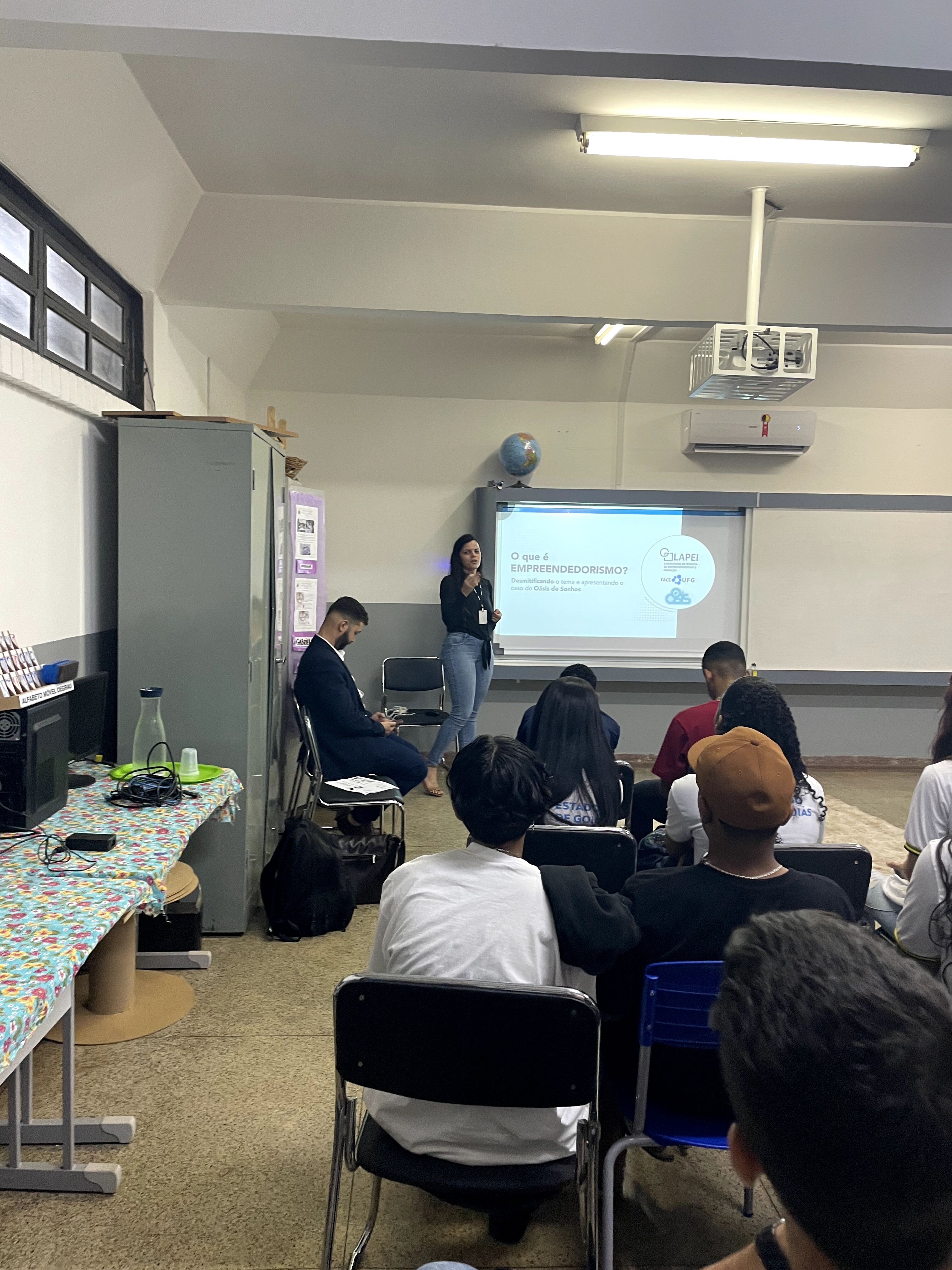 Foto palestra empreendedorismo na escola 04 - mulher morena, jovem, cabelos pretos, vestindo calça jeans e camisa preta, palestra para alunos do colégio presentes em uma sala de aula. 