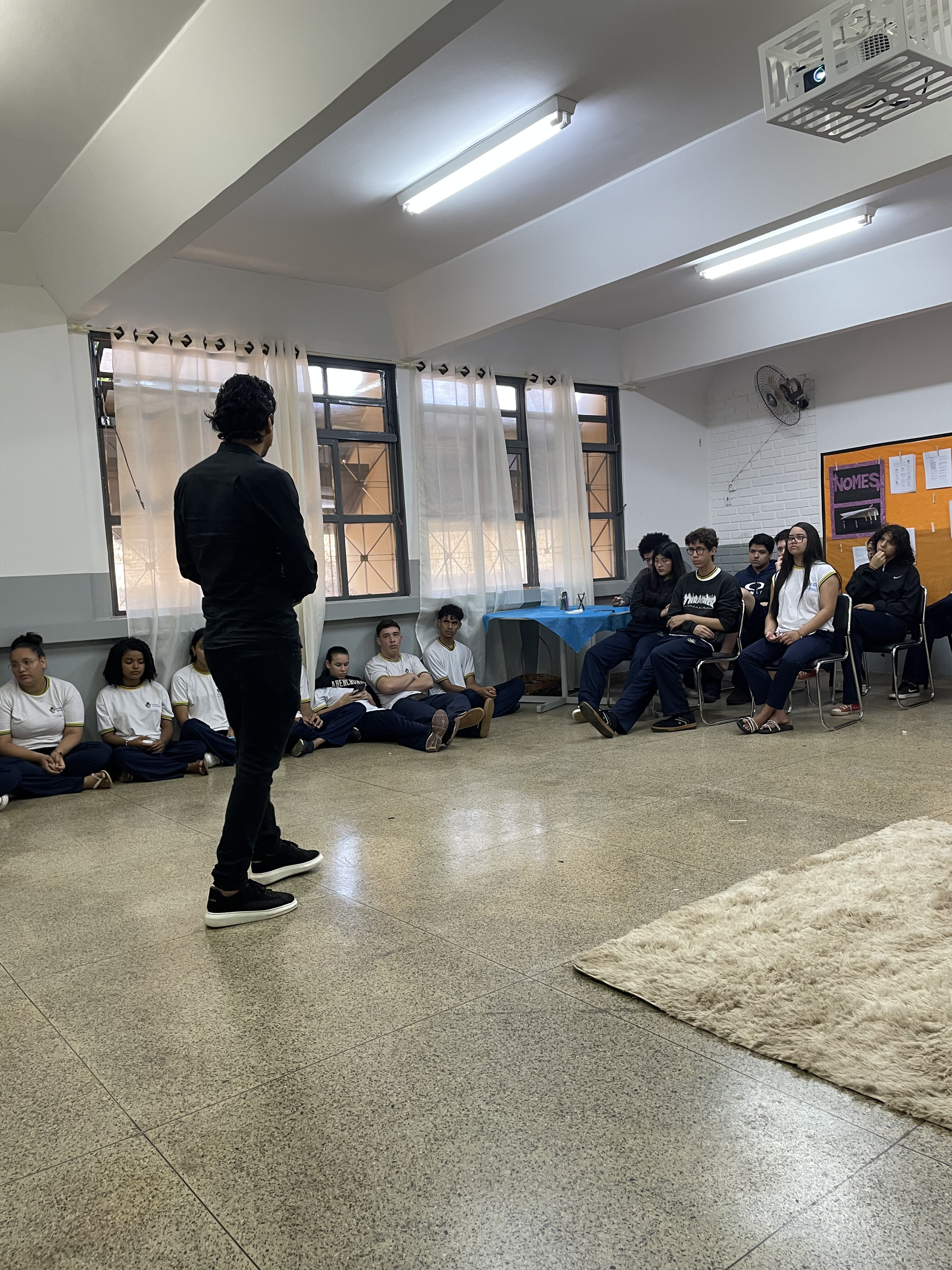 Foto palestra empreendedorismo na escola 05 - homem jovem, de costas, cabelo preto, vestindo calça preta, camisa preta, palestra para alunos do colégio, sentados ao redor de uma sala de aula. 