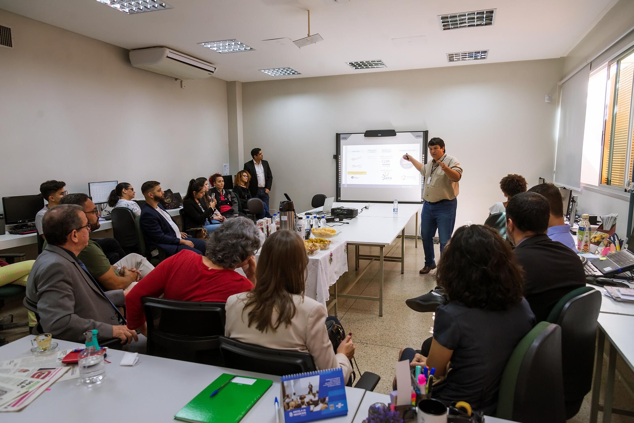 Nesta imagem aparece pessoas sentada dentro de uma sela de aula e outra pessoa em pé apresentando. 