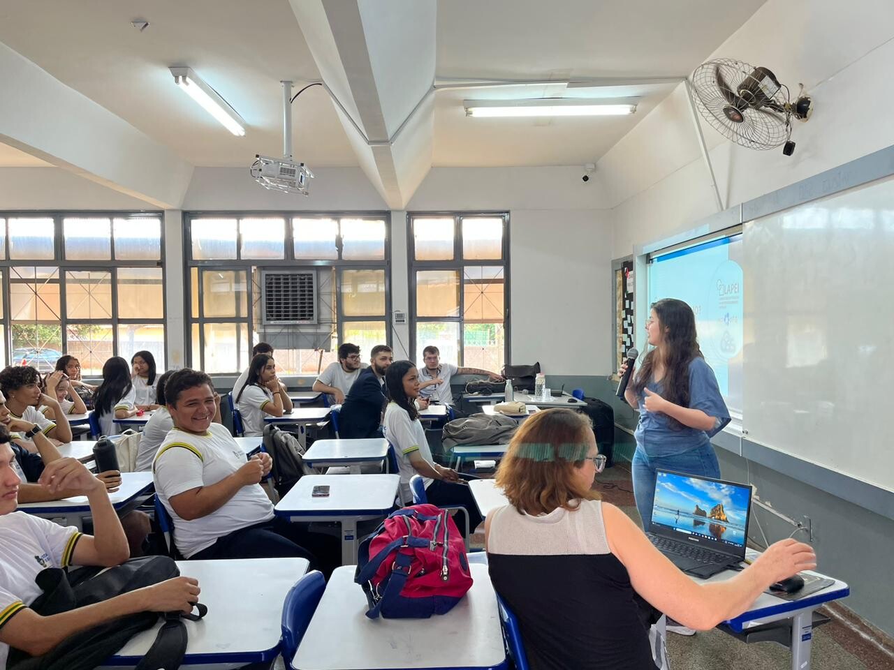 Foto palestra empreendedorismo na escola 02 - Mulher branca, jovem, vestindo calça jeans, blusa azul, de óculos, em pé e segurando um microfone, palestra para uma sala de aula cheia de alunos sentados.
