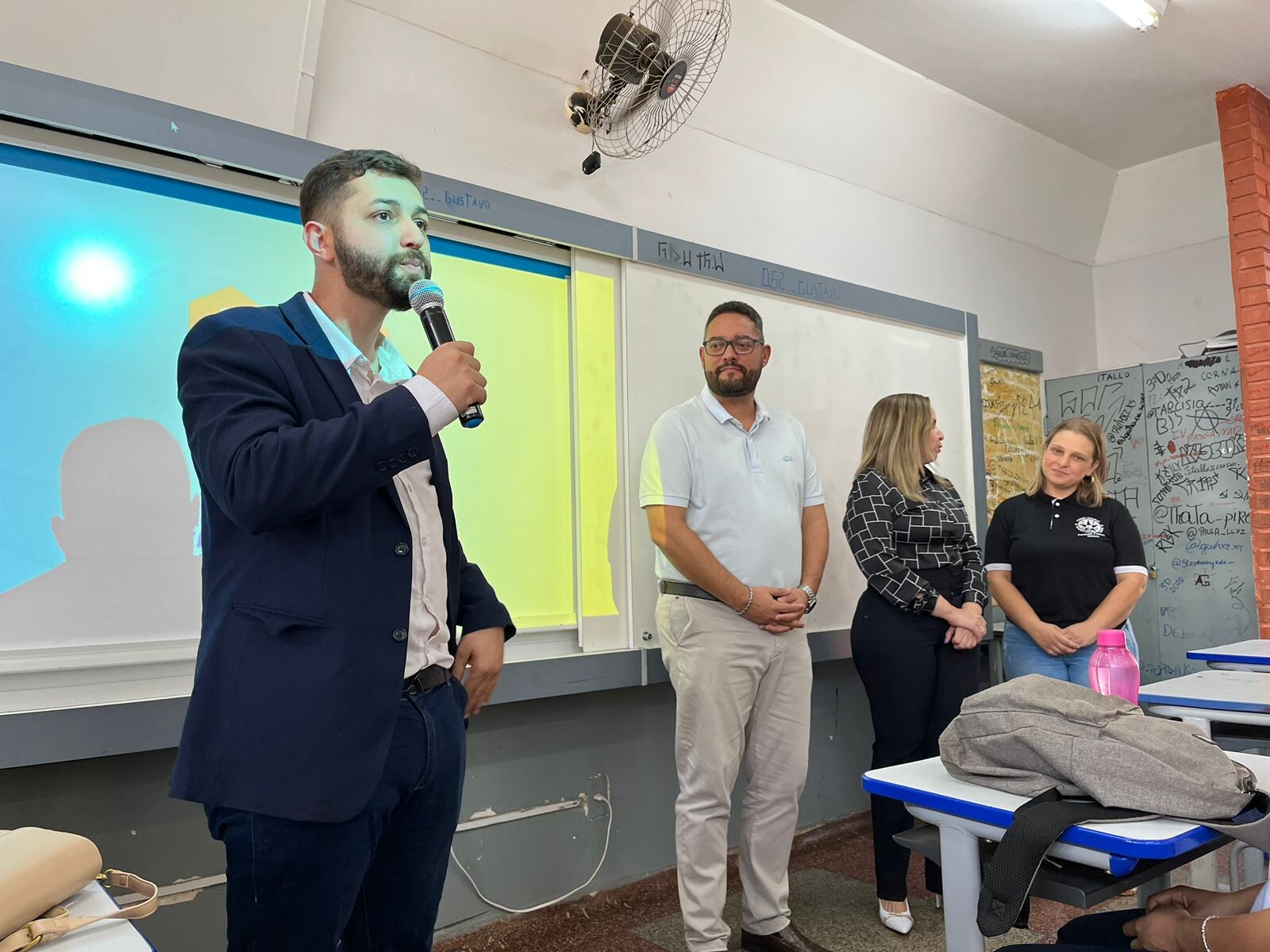 Foto palestra empreendedorismo na escola 01 - Homem branco, jovem, vestido de paletó preto, calça social preta, camisa branca, de pé, segurando um microfone. Ao fundo, um homem e duas mulheres de pé. 