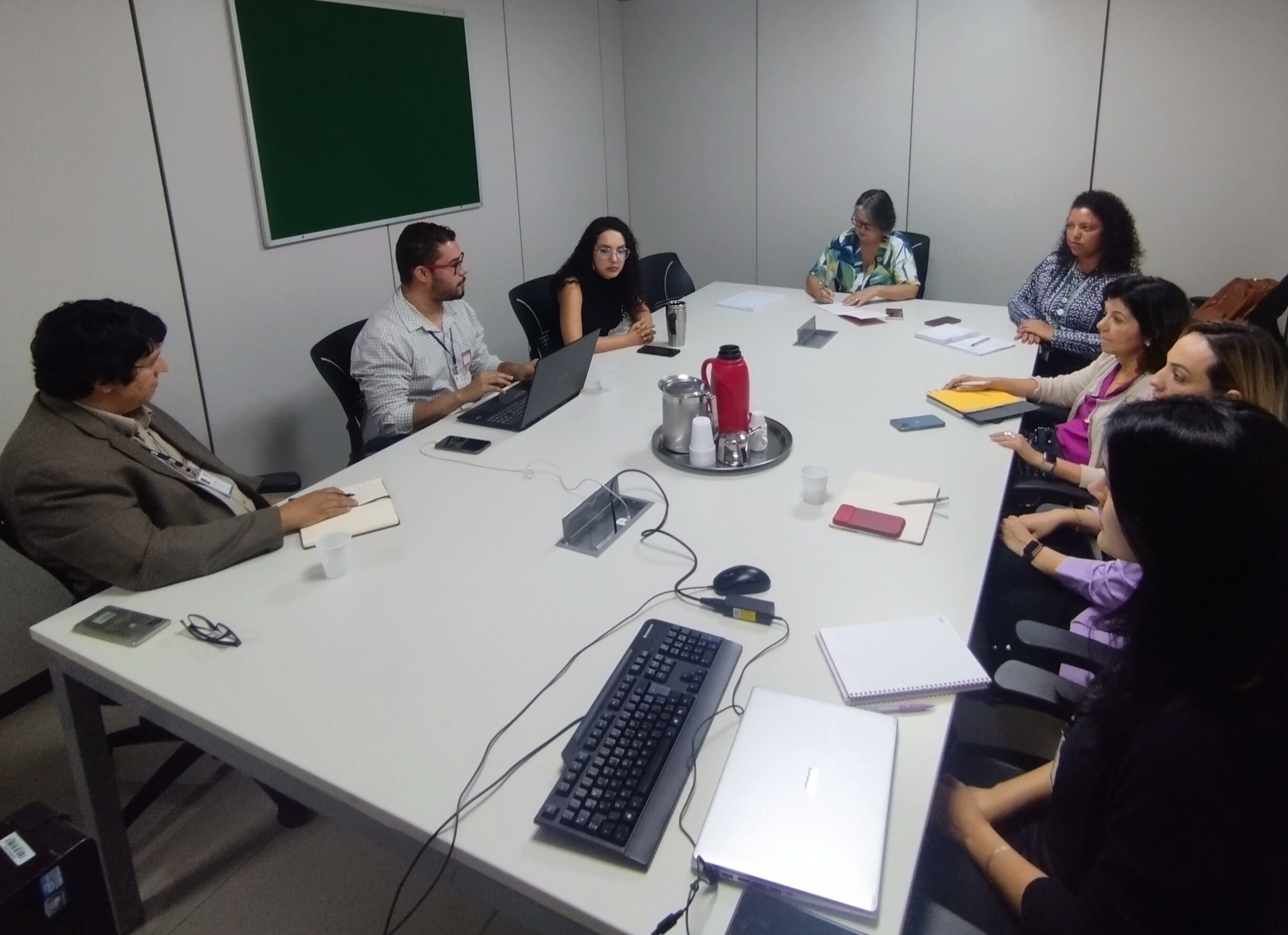 Na foto aparecem 8 pesquisadores, sentados ao redor de uma grande mesa cinza de reunião. Seis são mulheres e dois homens.