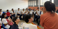 #ParaTodosVerem:

Fotografia de estudantes em uma sala de aula dispostos em círculo. Em primeiro plano e de costas, uma mulher de blusa alaranjada gesticula enquanto fala com os jovens. Ao redor, os alunos vestem uniforme escolar de camiseta branca com detalhes amarelos e azuis, sentados em cadeiras e sobre um tapete colorido no chão, todos atentos à conversa.