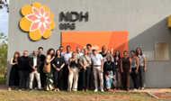 banner_notícia_visita_institucional_ppgdh
#ParaTodosVerem:

Fotografia em plano médio aproximado de um grupo de cerca de vinte pessoas alinhadas lado a lado e sorridentes, posando em frente ao edifício do NDH UFG.

Ao fundo, na parede cinza do prédio, destaca-se em grandes proporções a marca estilizada do NDH em formato de flor nos tons amarelo e rosa, ao lado do texto em relevo "NDH UFG" e de um painel laranja onde se lê parcialmente a palavra "escola". O grupo veste roupas casuais e formais em tons variados, como preto, bege, branco e estampado.