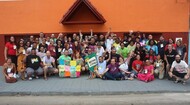 #ParaTodosVerem:

Fotografia em plano aberto de um grande grupo de pessoas reunidas ao ar livre, posando em frente a um edifício de fachada alaranjada com telhado triangular no centro.

As pessoas estão dispostas em várias fileiras — algumas sentadas no chão, outras agachadas e a maioria em pé ao fundo. Elas sorriem, e muitas acenam ou fazem gestos com as mãos. No centro, o grupo segura uma colcha colorida com retalhos e uma estandarte verde onde se lê "Rede Caminho e Esperança". Todos usam roupas casuais e crachás de identificação pendurados no pescoço.