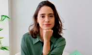 banner_notícia_semi-finalista
#ParaTodosVerem:

Retrato em primeiro plano de uma jovem de pele clara e cabelos castanhos curtos. Ela está ligeiramente inclinada para a frente, com o queixo apoiado na mão e um semblante sereno, olhando para a câmera. Veste uma camisa de cetim verde-oliva. Ao fundo, uma parede branca iluminada e, no canto esquerdo, folhas de uma planta verde. No canto inferior direito, aparece a ponta de um livro verde-claro.