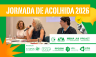 #ParaTodosVerem: Banner retangular com moldura verde. No topo, sobre uma larga faixa amarela, está escrito em letras brancas: "JORNADA DE ACOLHIDA 2026". No centro, há uma fotografia de três pessoas sentadas à mesa de um evento: à esquerda, uma mulher loira sorri; no centro, uma mulher de cabelos grisalhos fala ao microfone; e à direita, um homem de óculos observa. Sobreposta à direita da foto, há uma colagem com uma mão em preto e branco segurando a ilustração de uma lâmpada amarela acesa. No rodapé, sobre uma faixa branca, alinham-se em verde as logomarcas das instituições organizadoras e parceiras, como USP, UFG, Media Lab, entre outras.