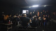 #ParaTodosVerem:

Fotografia em plano aberto de uma palestra em um galpão com paredes pretas e teto escuro, iluminado por lâmpadas no centro. Uma mulher de cabelos volumosos e grisalhos fala em pé, ao fundo, ao lado de uma tela de projeção ligada com o título "Mediar é ancestral".

A plateia está disposta em semicírculo em primeiro plano e vista de costas, sentada em bancos de madeira e cadeiras plásticas escuras. Nas paredes pretas do espaço, há crânios decorativos de bois pendurados.