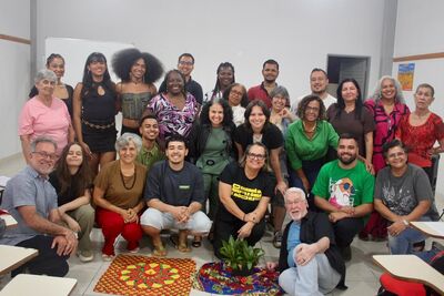 banner_noticia_cafe_com_direitos_humanos

#ParaTodosVerem Uma fotografia em grupo, colorida, de cerca de 25 pessoas de diversas idades, etnias e origens. Elas estão reunidas em quatro fileiras, algumas agachadas na frente, outras de pé atrás, sorrindo e olhando para a câmera. Estão num espaço interior amplo, que parece ser uma sala de aula ou um auditório, com paredes brancas, portas cinzentas e o que parecem ser mesas de formica visíveis nas bordas inferiores da imagem. No chão, à frente do grupo, há dois panos coloridos e estampados sobre um tapete menor e circular, com uma pequena planta em vaso no centro. As pessoas vestem uma variedade de roupas casuais e coloridas, incluindo t-shirts, blusas, vestidos e calças. Alguns têm óculos e chapéus. A iluminação é natural e difusa. A atmosfera geral é de calor e camaradagem.