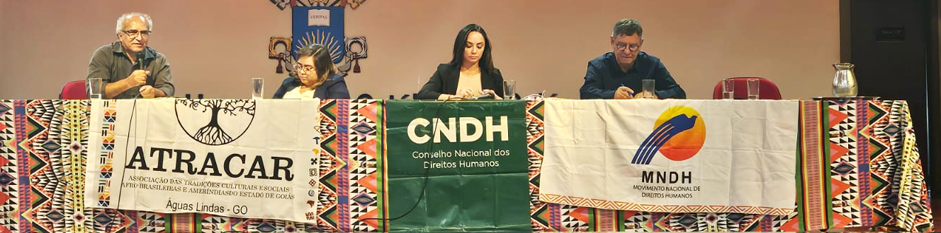 #PraTodosVerem: Fotografia de quatro pessoas sentadas lado a lado atrás de uma mesa de debate em um evento. Da esquerda para a direita, há um homem grisalho falando ao microfone, seguido por duas mulheres e outro homem grisalho que leem ou fazem anotações. A mesa está coberta por um tecido com estampas geométricas coloridas e exibe três faixas institucionais penduradas na frente: ATRACAR (à esquerda), CNDH (ao centro, em verde) e MNDH (à direita). Ao fundo, uma parede clara.