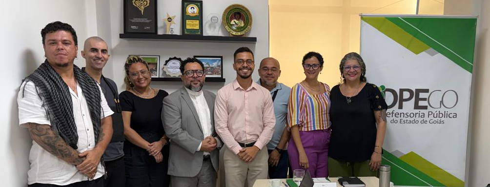 Banner_Topo_Notícia_CPDR - #ParaTodosVerem: Fotografia colorida de um grupo de oito pessoas em pé, sorrindo, em pose formal dentro de um escritório bem iluminado. Ao fundo, há duas prateleiras pretas fixadas na parede com diversos prêmios e troféus. À direita, um banner corporativo de roll-up verde e branco exibe o logotipo "DPE-GO" e o texto "Defensoria Pública do Estado de Goiás". As pessoas, que incluem homens e mulheres, vestem trajes variados, do casual ao profissional.