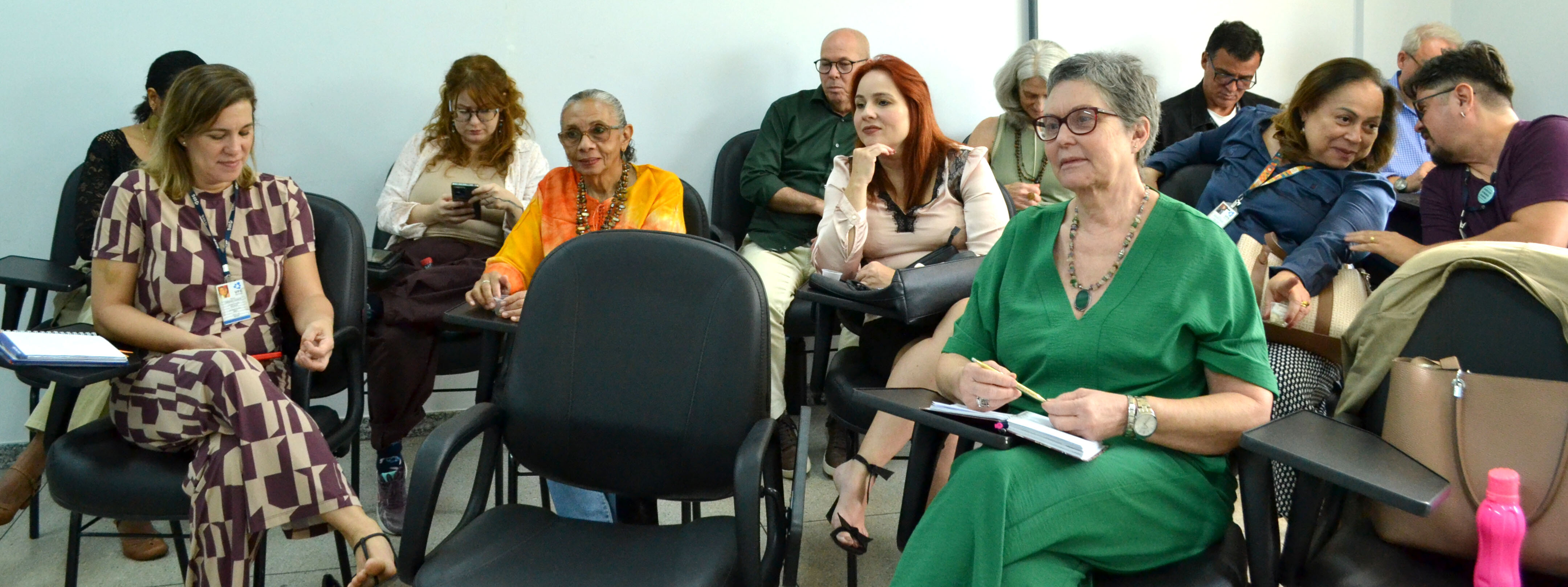 visita_institucional_docentes#ParaTodosVerem:Grupo de pessoas sentadas em cadeiras universitárias prestando atenção a uma apresentação. Em primeiro plano, à direita, destaca-se uma mulher de cabelo curto grisalho e óculos, vestindo um conjunto verde, com caderno e caneta nas mãos. À esquerda, em primeiro plano, uma mulher veste um fato estampado geométrico em bege e castanho. Nas filas de trás, observam atentamente outros participantes, incluindo uma mulher de túnica alaranjada, outra de blusa rosa-claro e homens em conversa discreta ao fundo.