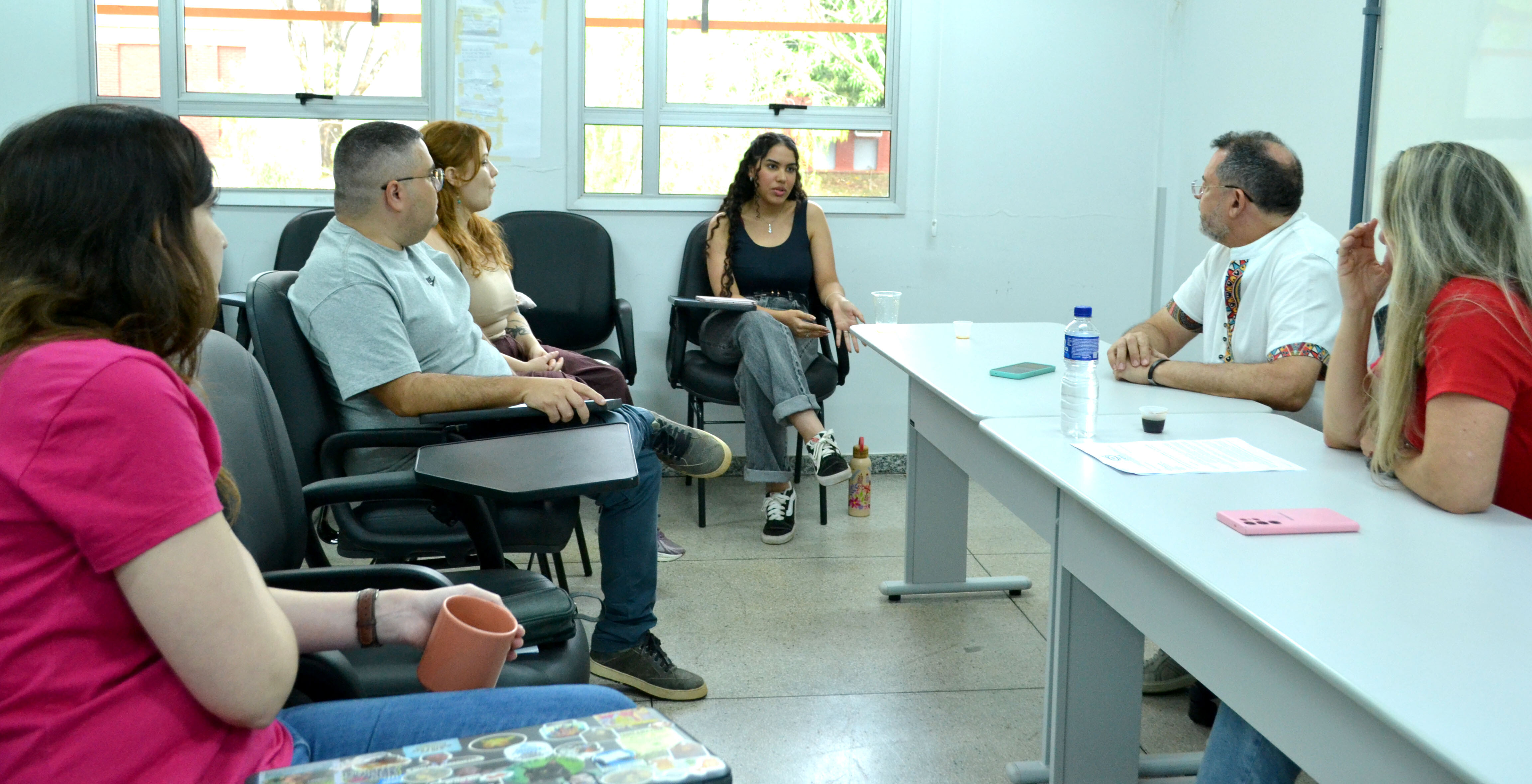 Reunião da Coordenação do PPGDH#ParaTodosVerem:Fotografia em plano médio de uma reunião numa sala iluminada. À esquerda, pessoas sentadas em cadeiras universitárias escutam atentamente, incluindo uma mulher em primeiro plano de t shirt rosa-choque a segurar uma caneca, um homem de t shirt cinzenta e calças jeans, e uma mulher ruiva. Ao fundo, ao centro, uma jovem de regata preta e calças jeans fala gesticulando. À direita, à mesa cinzenta com uma garrafa de água e telemóveis, estão sentados um homem de bata branca e uma mulher de t shirt vermelha.