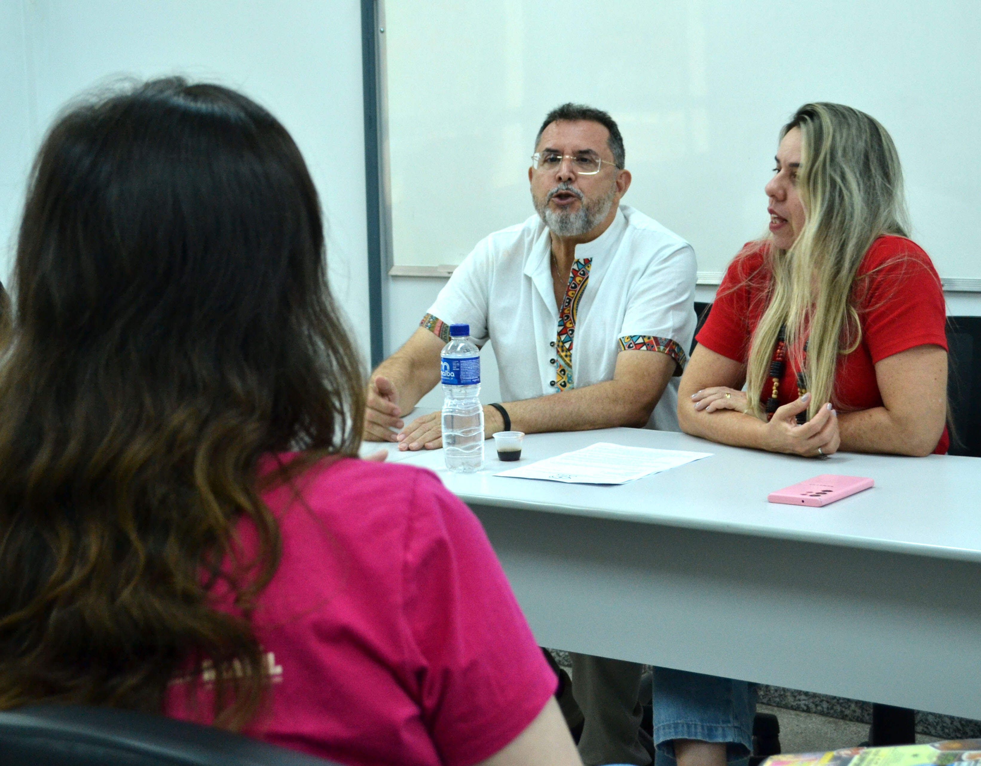 visita_intitucional_coordenação_ppgdh#ParaTodosVerem:Fotografia em plano médio focada em duas pessoas sentadas a uma mesa branca de reunião. Ao centro, um homem de óculos, barba e camisa branca com detalhes étnicos coloridos na gola e mangas fala gesticulando. À sua direita, uma mulher de cabelos loiros compridos e t-shirt vermelha escuta e intervém na conversa. Em primeiro plano, de costas para a câmara e com o rosto oculto, vê-se a cabeça e os ombros de uma mulher de t-shirt rosa-choque. Sobre a mesa, estão uma garrafa de água, papéis e um telemóvel rosa.