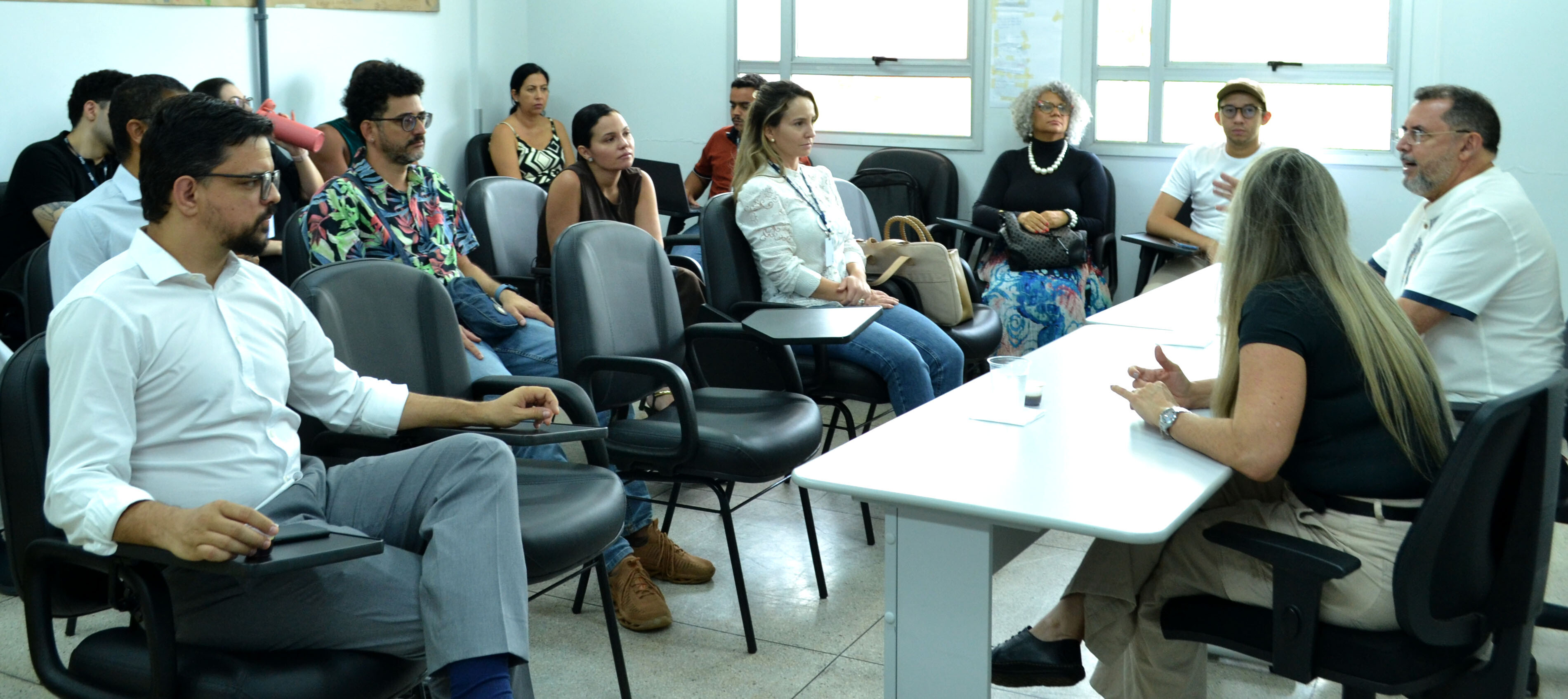 visita_institucional_discentes#ParaTodosVerem:Visão geral de uma reunião em sala de aula. À esquerda, participantes atentos sentam-se em cadeiras estofadas, com destaque para um homem de camisa social branca em primeiro plano e outro de camisa florida ao lado. À direita, numa mesa branca, uma mulher loira de blusa preta gesticula ao falar, enquanto um homem de bata branca ao fundo também intervém. Ao fundo, outros participantes observam junto às janelas.