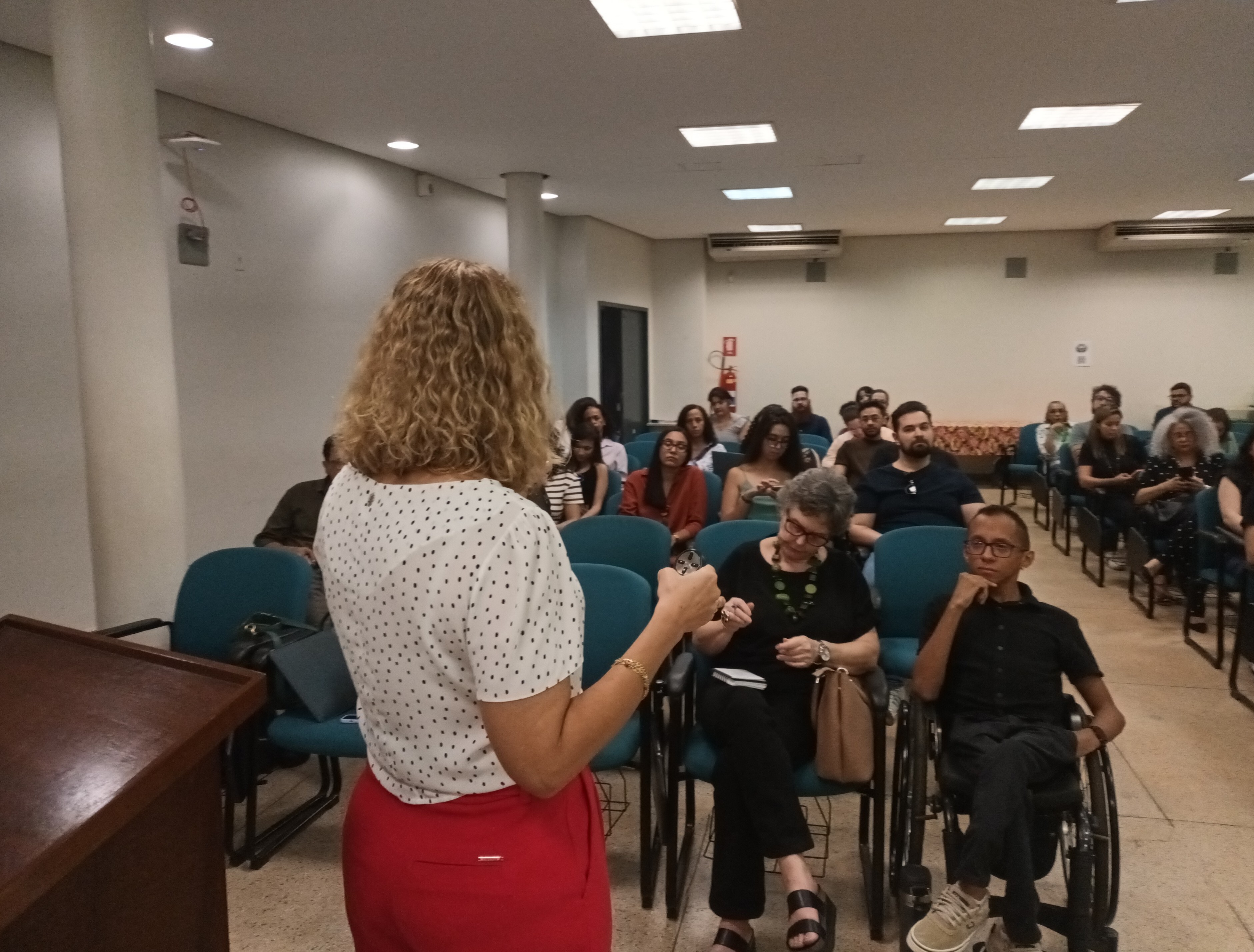 #ParaTodosVeremFotografia colorida em plano médio, tirada de costas para a palestrante em direção à plateia. No primeiro plano, à esquerda, vemos parte do corpo de uma mulher de cabelos loiros cacheados, vestindo uma blusa branca com bolinhas pretas e calça vermelha, de frente para o público.A plateia é composta por cerca de vinte pessoas sentadas em poltronas azuis, organizadas em fileiras. Na primeira fila, em destaque, uma mulher de cabelos grisalhos e colar verde observa algo em suas mãos, e ao lado dela, à direita, um homem de pele parda e óculos, sentado em uma cadeira de rodas, olha atentamente para a palestrante. O ambiente é um auditório com iluminação embutida no teto, paredes claras e aparelhos de ar-condicionado ao fundo.
