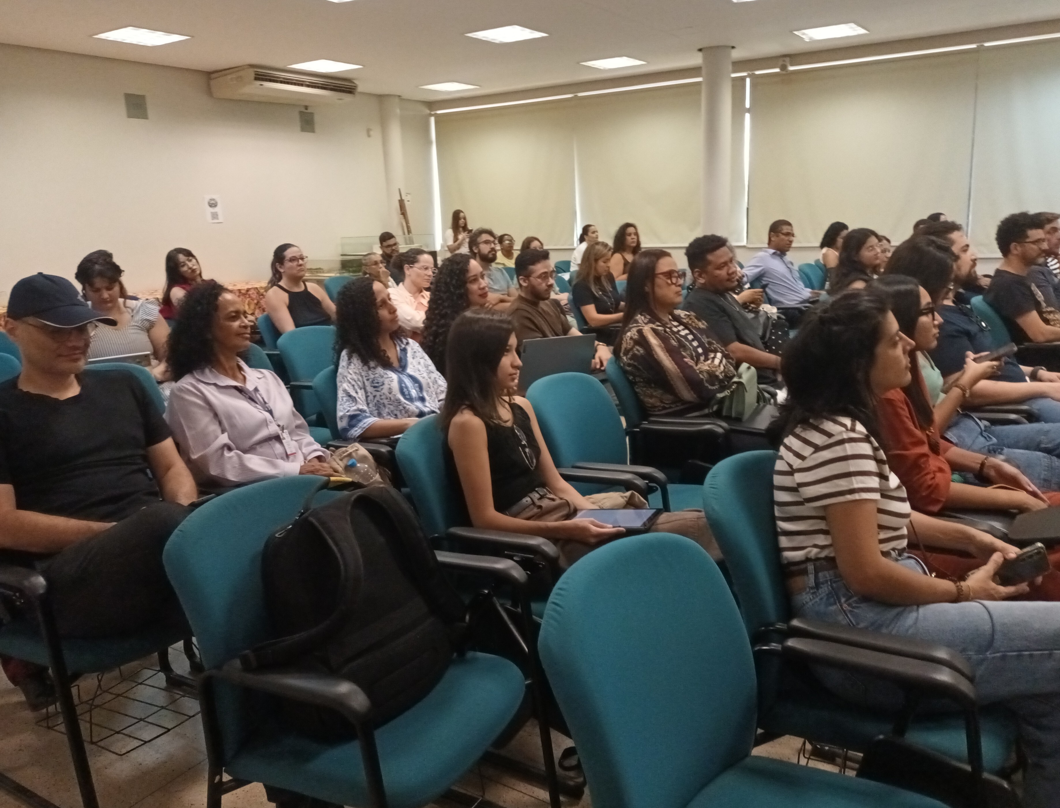 #ParaTodosVeremFotografia colorida em plano médio e ângulo lateral da plateia em um auditório. Homens e mulheres de diferentes idades e etnias estão sentados em poltronas azuis, dispostas em fileiras.Em primeiro plano, à esquerda, um homem de boné e camiseta preta e, ao lado dele, uma mulher de camisa lilás. À direita, uma mulher de blusa listrada segura um celular. Ao fundo, as demais pessoas observam atentamente a palestra. O ambiente possui paredes claras, janelas com persianas beges fechadas ao fundo e iluminação embutida no teto. Uma mochila preta está apoiada em uma das poltronas vazias à esquerda.