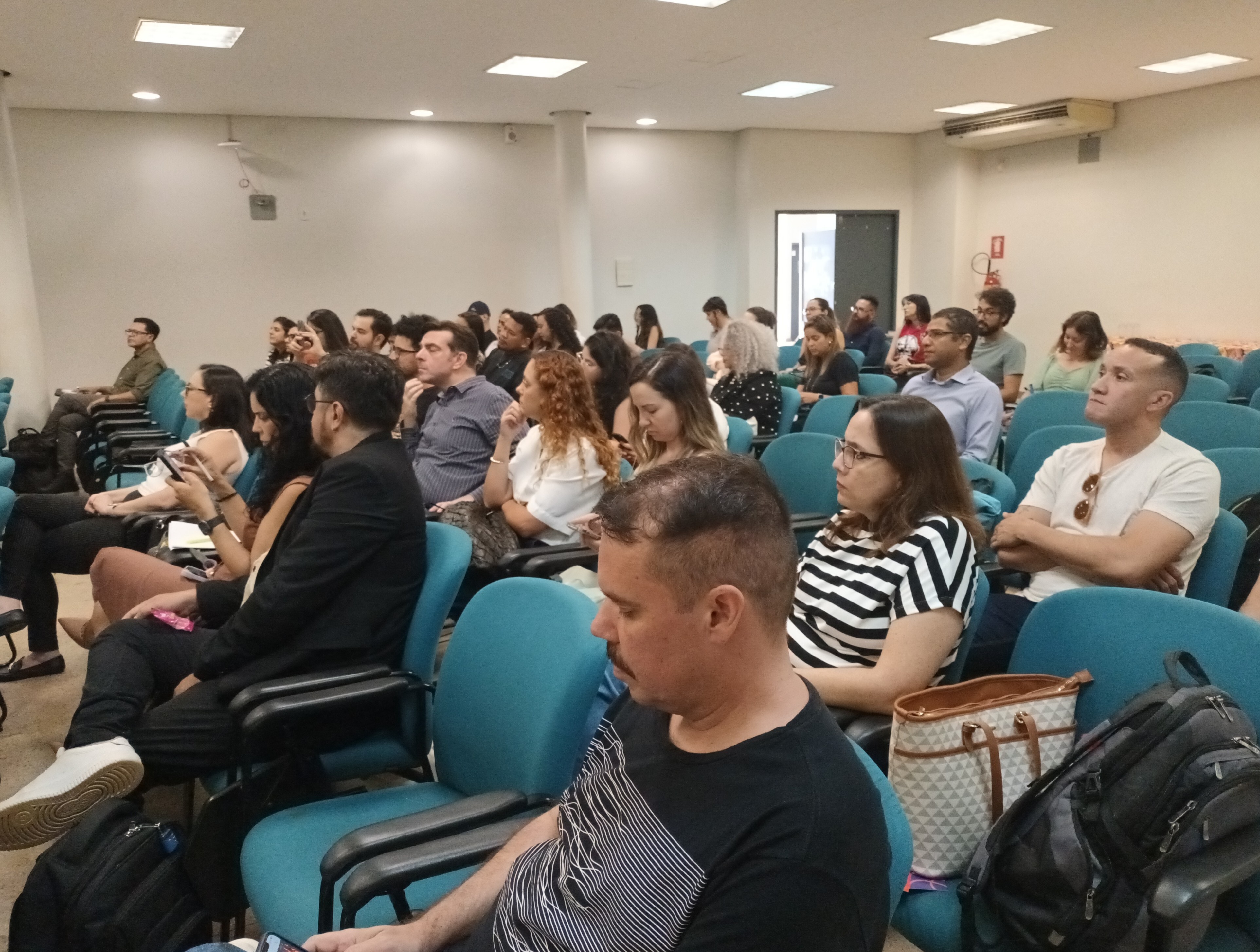 #ParaTodosVeremFotografia colorida em plano médio e ângulo lateral da plateia em um auditório. Diversas pessoas de perfis variados estão sentadas em poltronas estofadas na cor azul, dispostas em fileiras que preenchem o ambiente.Em primeiro plano, à direita, um homem de camiseta preta com estampas de linhas brancas e, ao lado dele, uma mulher com blusa listrada em preto e branco. Os demais participantes, ao fundo, aparecem sentados e voltados para a esquerda, sugerindo que acompanham uma apresentação. O auditório tem paredes claras, piso de granilite cinza e teto com luminárias quadradas embutidas.