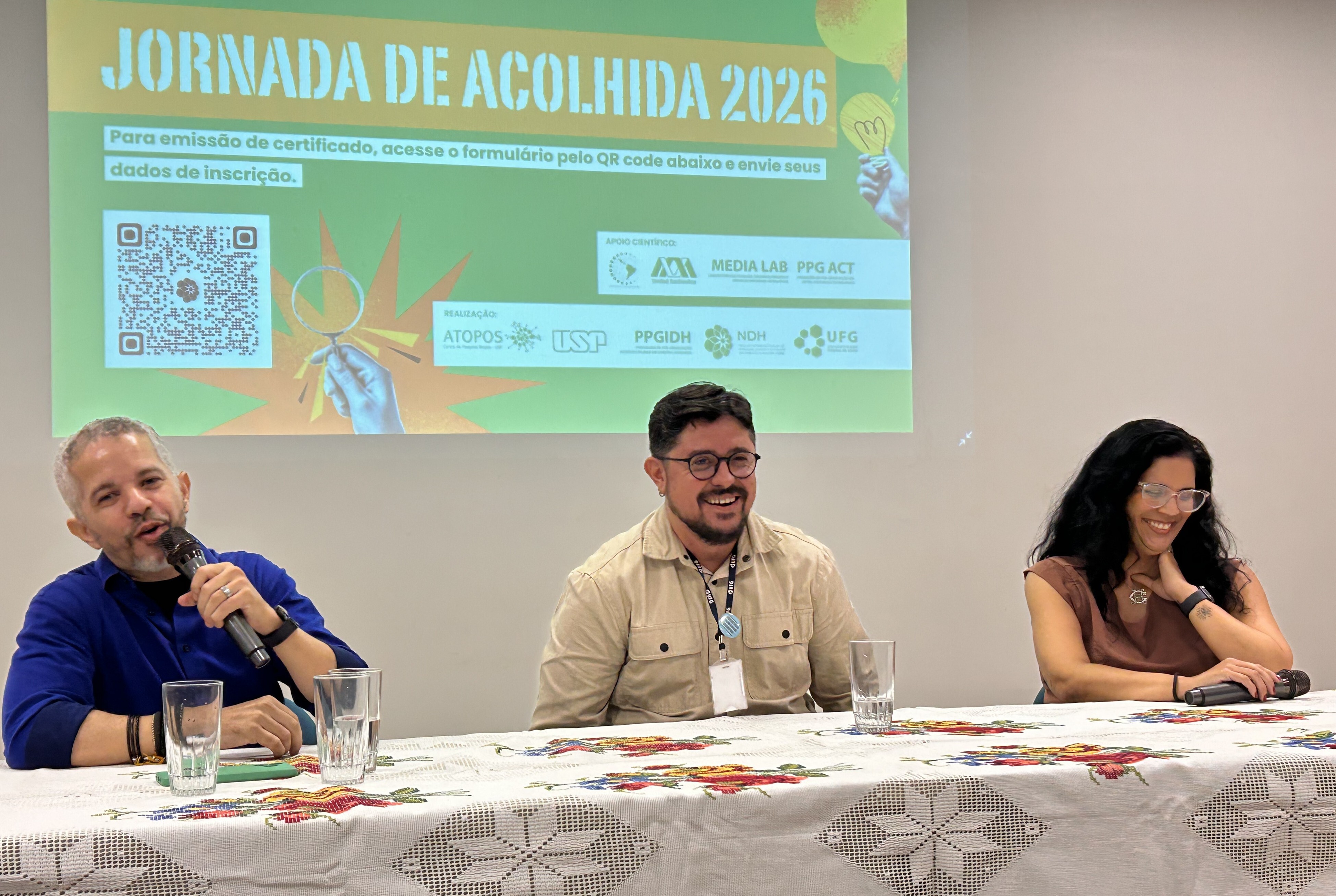 #ParaTodosVeremFotografia colorida em plano médio de três pessoas em uma mesa de debate. À esquerda, um homem de pele clara e barba rala, vestindo camisa azul-royal, fala ao microfone. Ao centro, um homem de pele clara, óculos e barba, vestindo uma camisa bege, sorri para o público. À direita, uma mulher de pele clara e longos cabelos pretos, usando óculos e blusa marrom, sorri com a mão no pescoço.Eles estão sentados atrás de uma mesa com toalha branca bordada com flores coloridas. Ao fundo, uma projeção em tons de verde e amarelo exibe o texto: "JORNADA DE ACOLHIDA 2026", acompanhado de um QR Code e logotipos de instituições como USP e UFG. O ambiente é bem iluminado e as paredes são brancas.