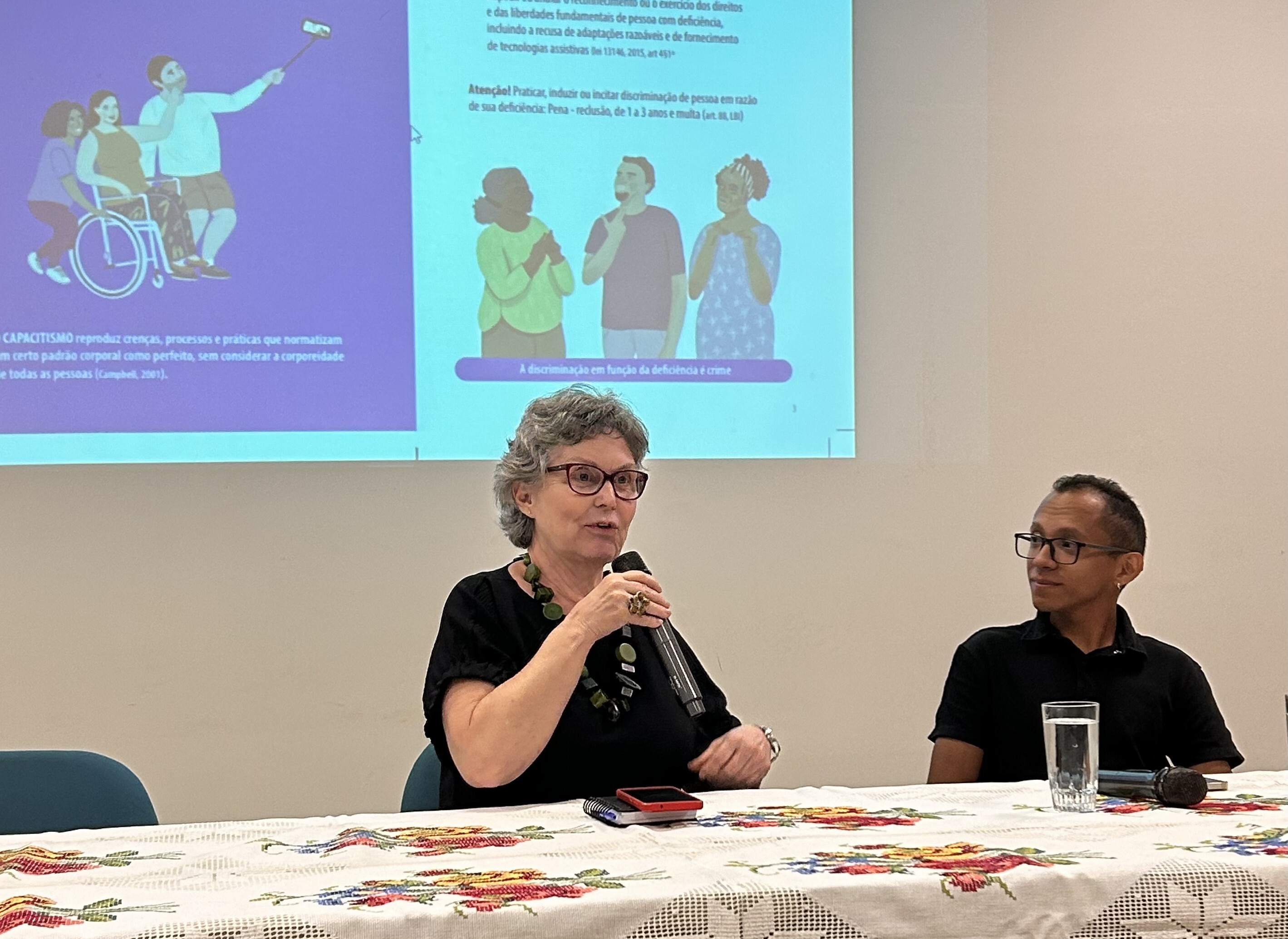#ParaTodosVeremFotografia colorida em plano médio de dois palestrantes sentados à frente de uma projeção. À esquerda, uma mulher de pele clara, cabelos grisalhos curtos e óculos de armação escura fala ao microfone. Ela usa uma blusa preta e um colar de pedras verdes. À direita, um homem de pele parda, cabelos curtos e óculos de grau observa a colega. Ele veste uma camisa polo preta.À frente deles, a mesa está coberta por uma toalha branca com bordados florais. Ao fundo, a projeção exibe um slide sobre o combate ao capacitismo, com ilustrações de pessoas com deficiência e textos explicativos em tons de roxo e azul. O ambiente possui iluminação clara e paredes brancas.