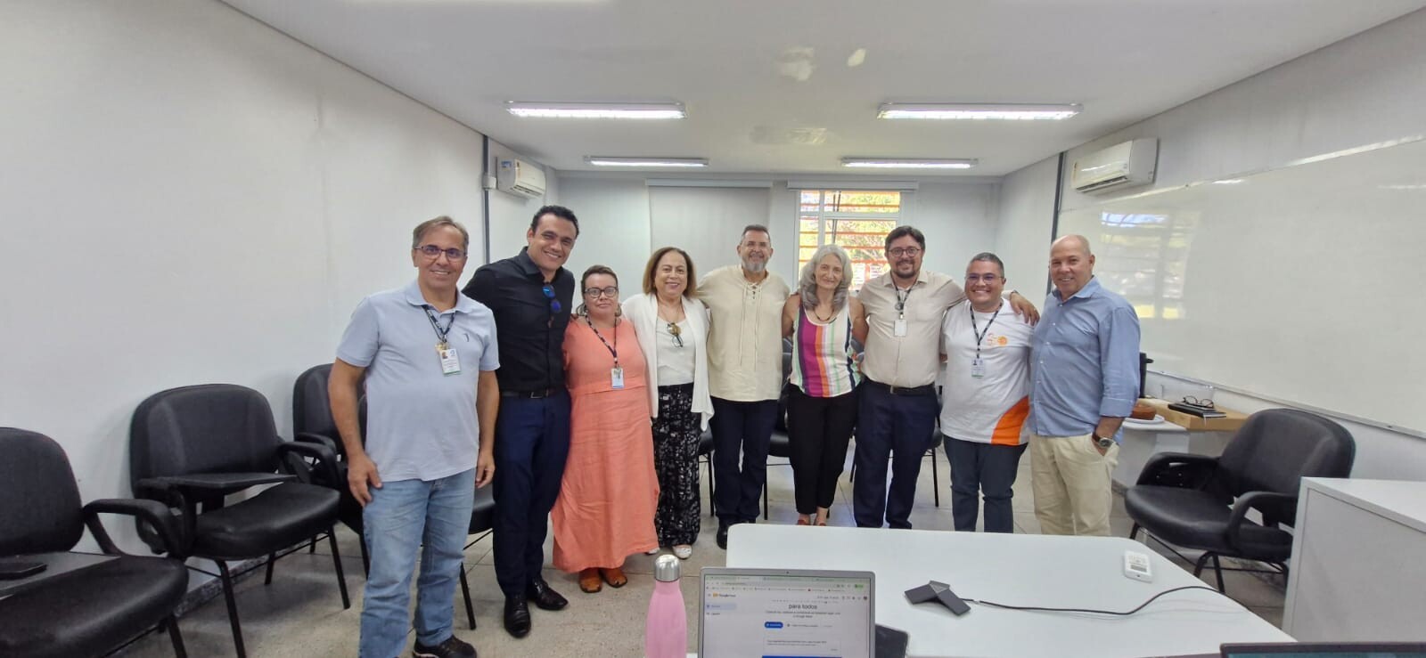 #ParaTodosVerem: Fotografia em plano aberto, em ambiente interno com iluminação clara, mostrando um grupo de nove pessoas alinhadas lado a lado, sorrindo para a câmera. O ambiente parece ser uma sala de reuniões ou de aula, com paredes brancas, cadeiras pretas de escritório ao fundo, um quadro branco à direita e uma mesa em primeiro plano na qual aparece o topo de um notebook aberto e uma garrafa rosa.